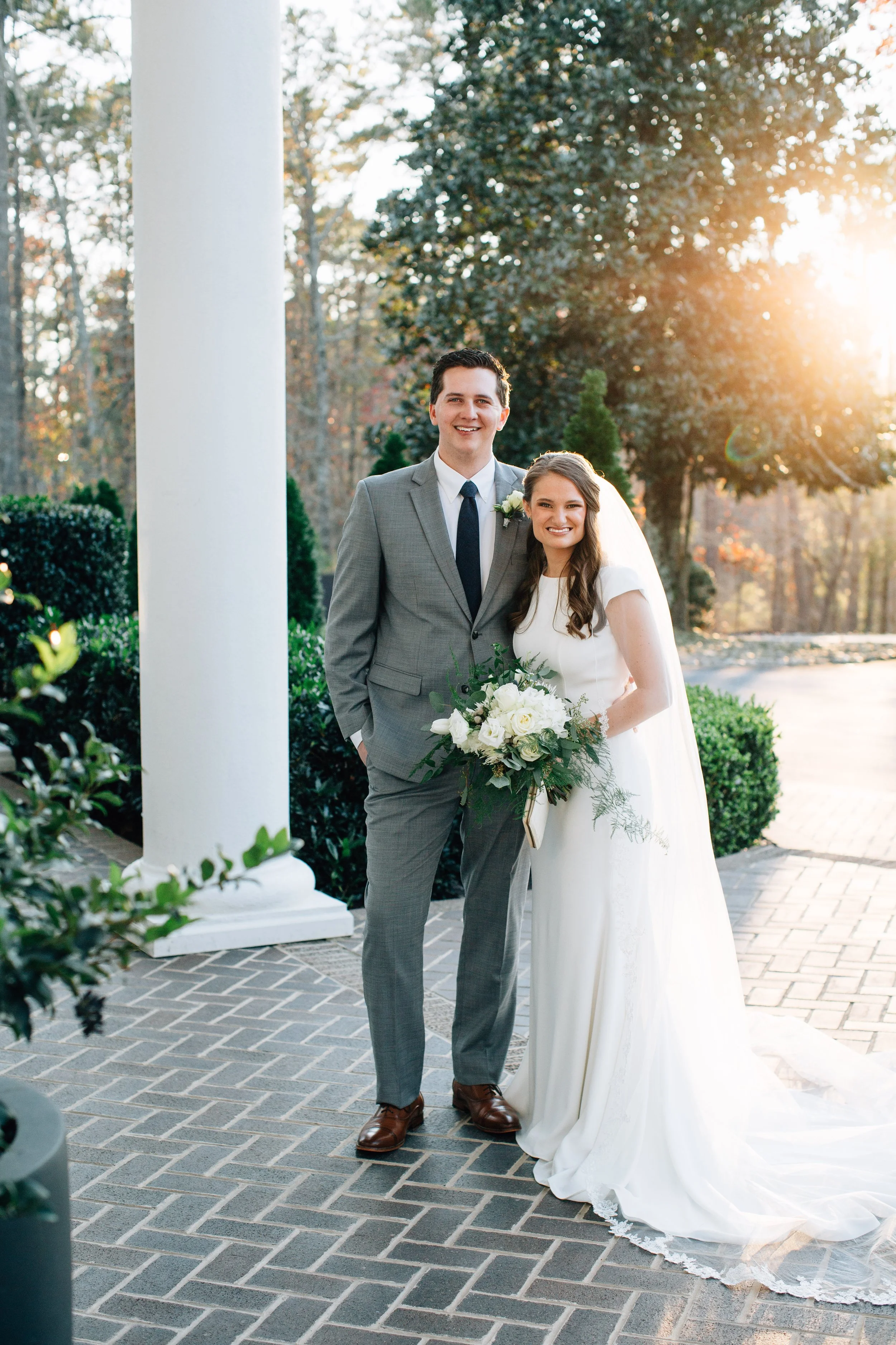A newlywed couple standing outdoors on a brick pathway during sunset, with greenery and trees in the background. The bride is in a white wedding gown holding a bouquet, and the groom is in a grey suit with a tie. They are smiling and posing for a wedding photo.