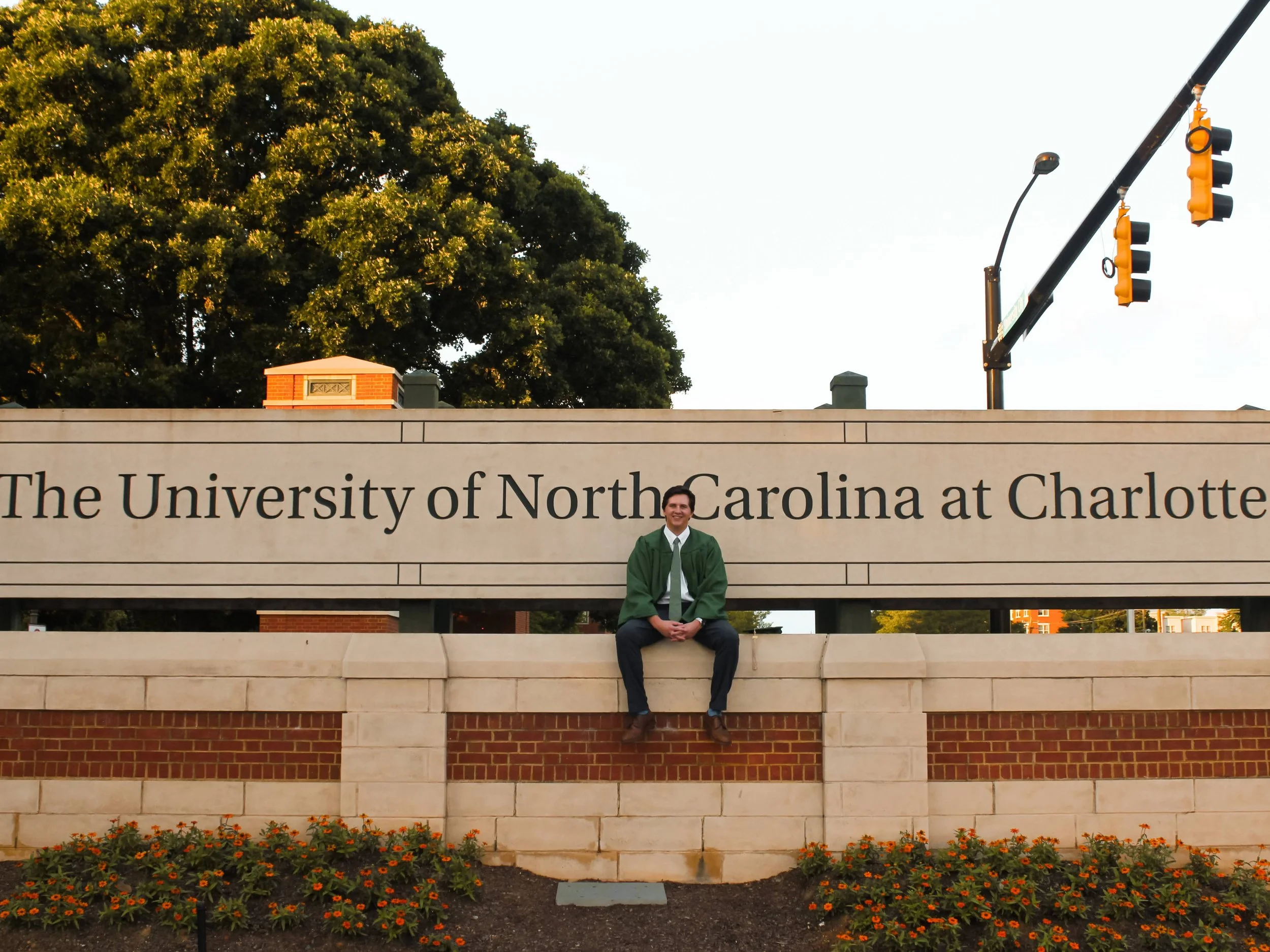 A person sitting on a stone ledge in front of a sign that reads 'The University of North Carolina at Charlotte' with trees and traffic lights in the background.