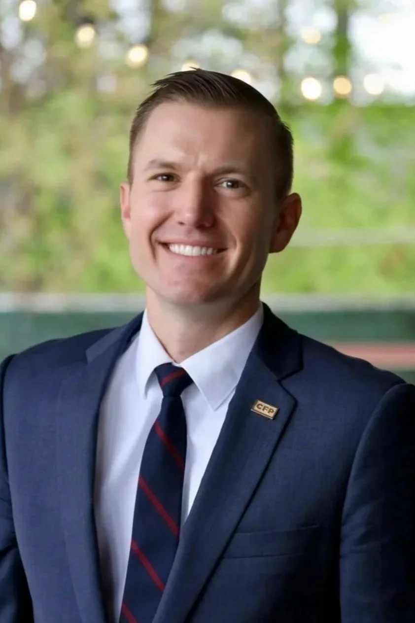 Portrait of a smiling man in a navy suit, white shirt, and striped tie, with a small gold lapel pin reading 'CFP', standing indoors with blurred greenery in the background.