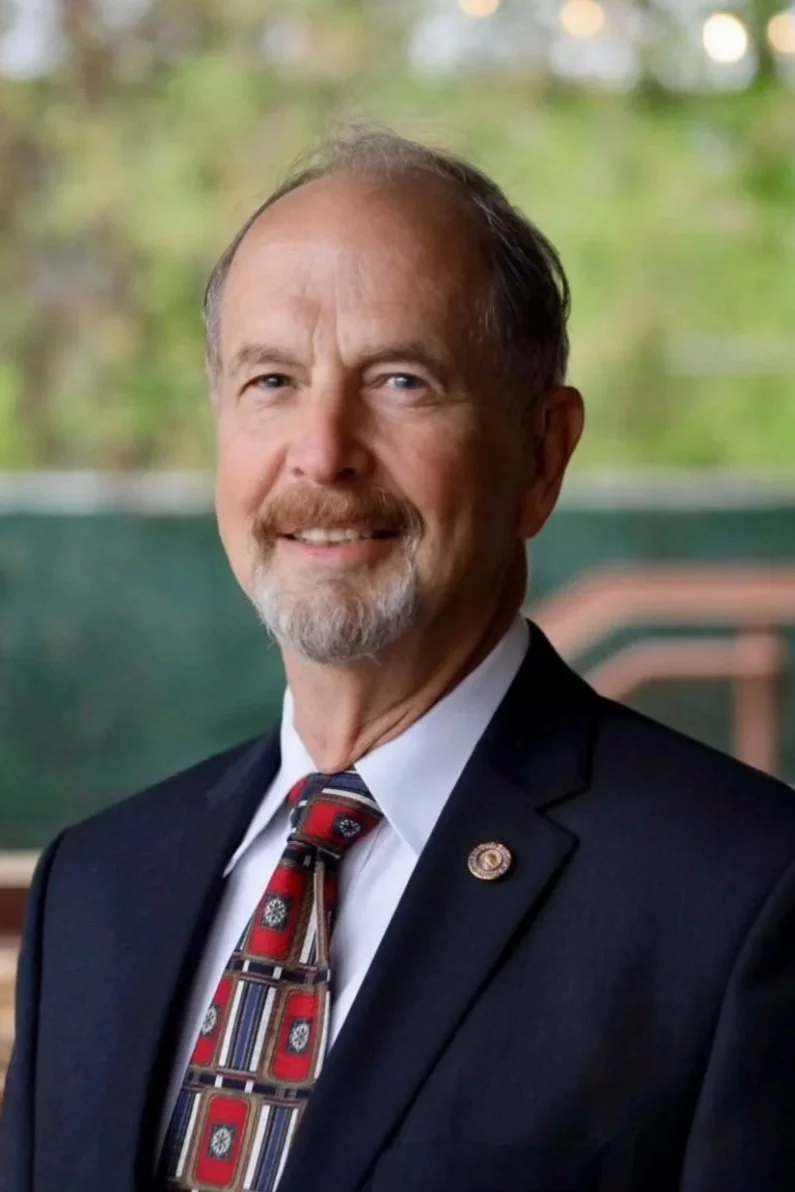 Portrait of a middle-aged man wearing a dark suit, white shirt, and a colorful patterned tie, smiling in front of a blurred outdoor background.