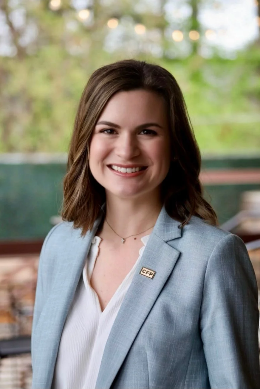A smiling woman with shoulder-length brown hair in a light blue blazer and white blouse, standing outdoors with blurred greenery in the background.