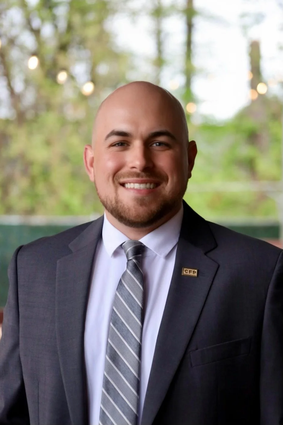 A man in a dark suit with a pin that says 'CFP' is smiling, with a blurred outdoor background with trees and string lights.