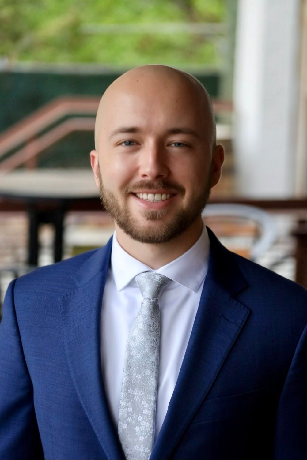 A smiling man in a blue suit, white shirt, and silver tie, standing indoors with a blurred background of a staircase and greenery.