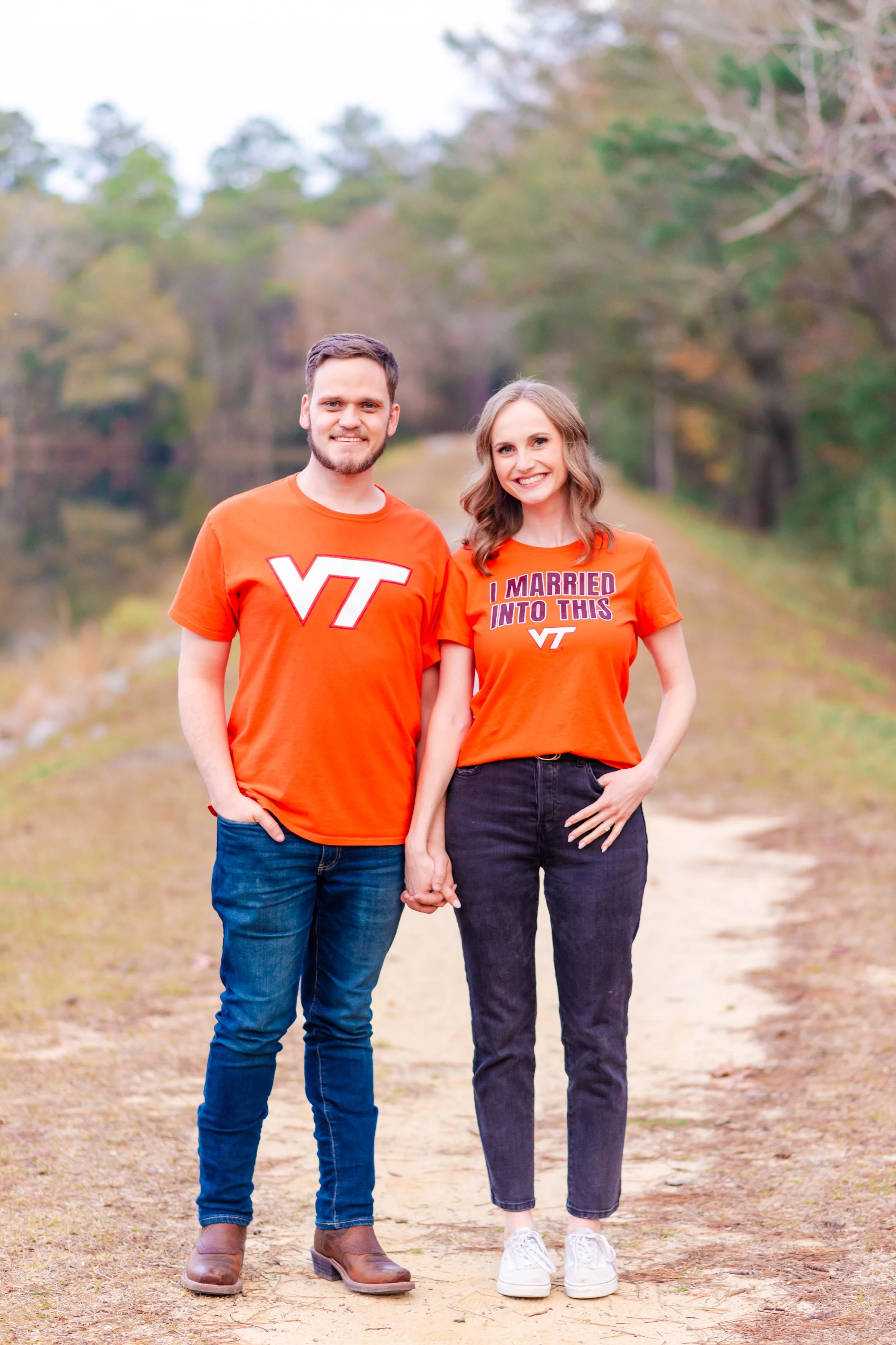 A happy couple holding hands and standing outdoors on a dirt path with trees in the background. They are both wearing orange shirts, with the man's shirt featuring the Virginia Tech logo and the woman's shirt reading 'I MARRIED INTO THIS VT.' The man has short brown hair and a beard, while the woman has wavy brown hair.