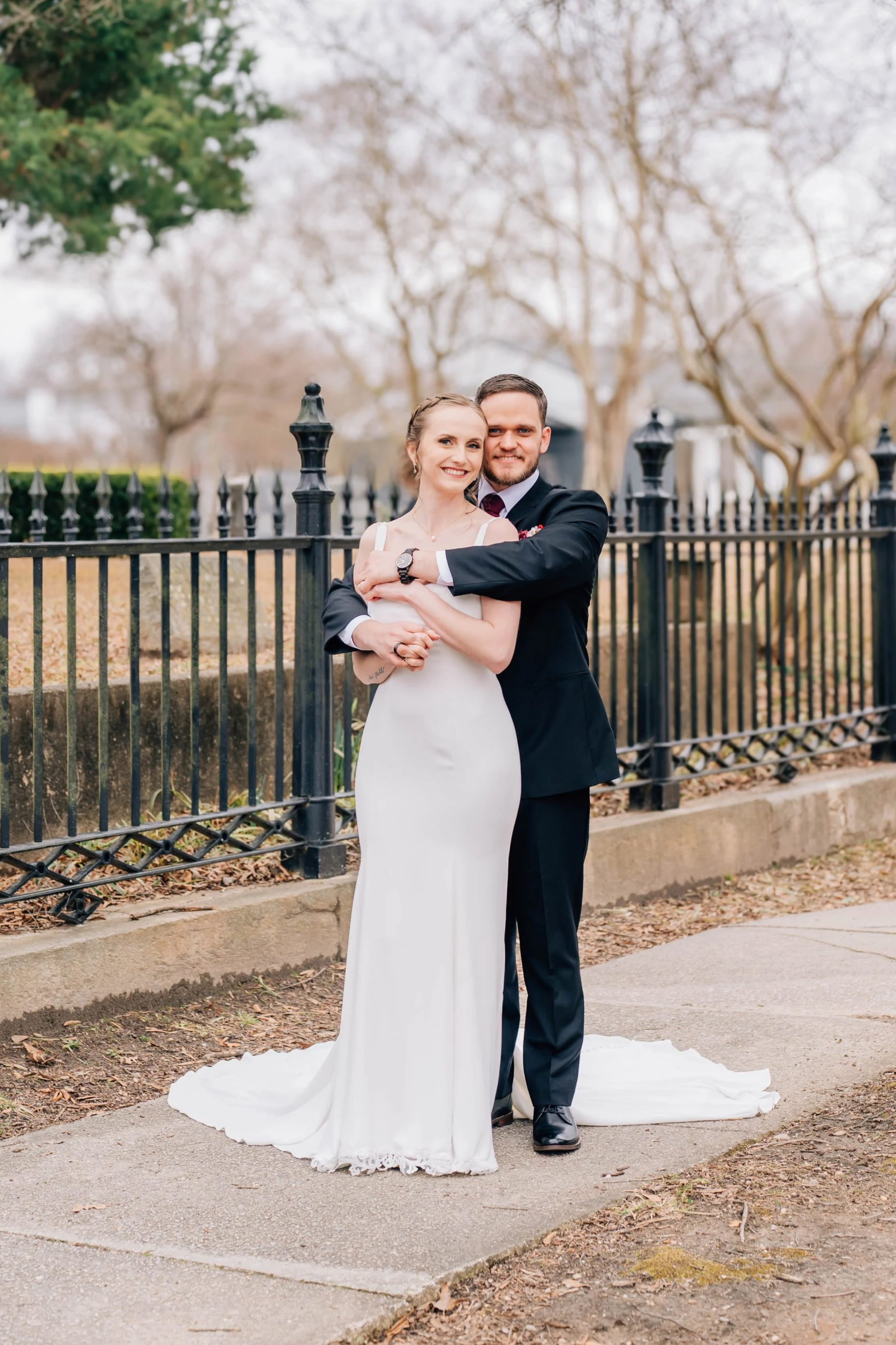 A bride and groom standing close together outdoors, hugging each other in front of a black wrought-iron fence. The bride is in a white wedding dress, and the groom is in a black suit. They are smiling and holding hands.
