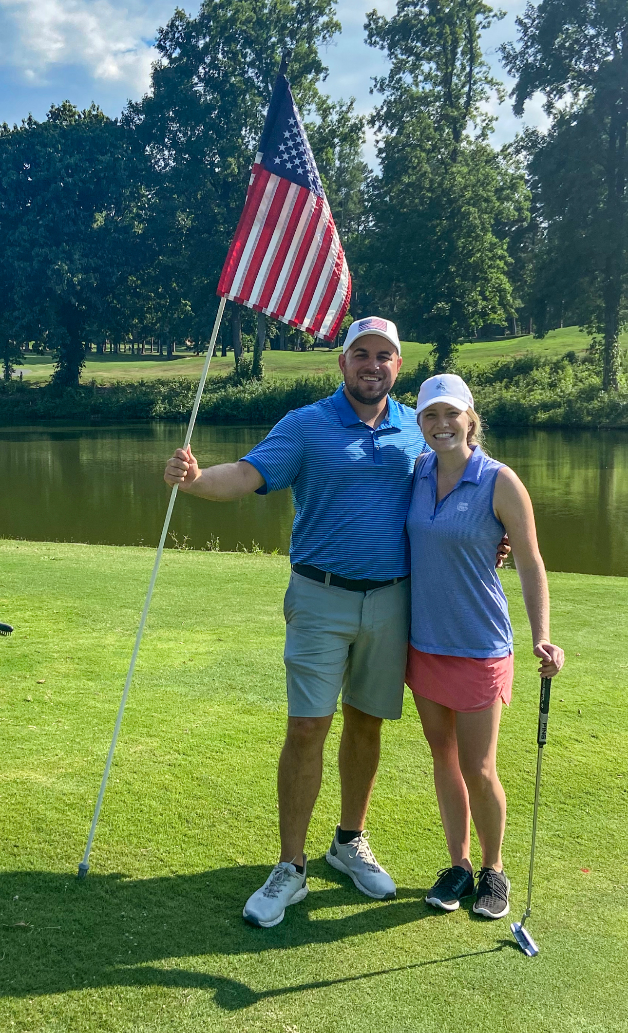 A man and a woman standing on a golf course, smiling at the camera. The man is holding an American flag and they are dressed in golf attire, with a pond and trees in the background.