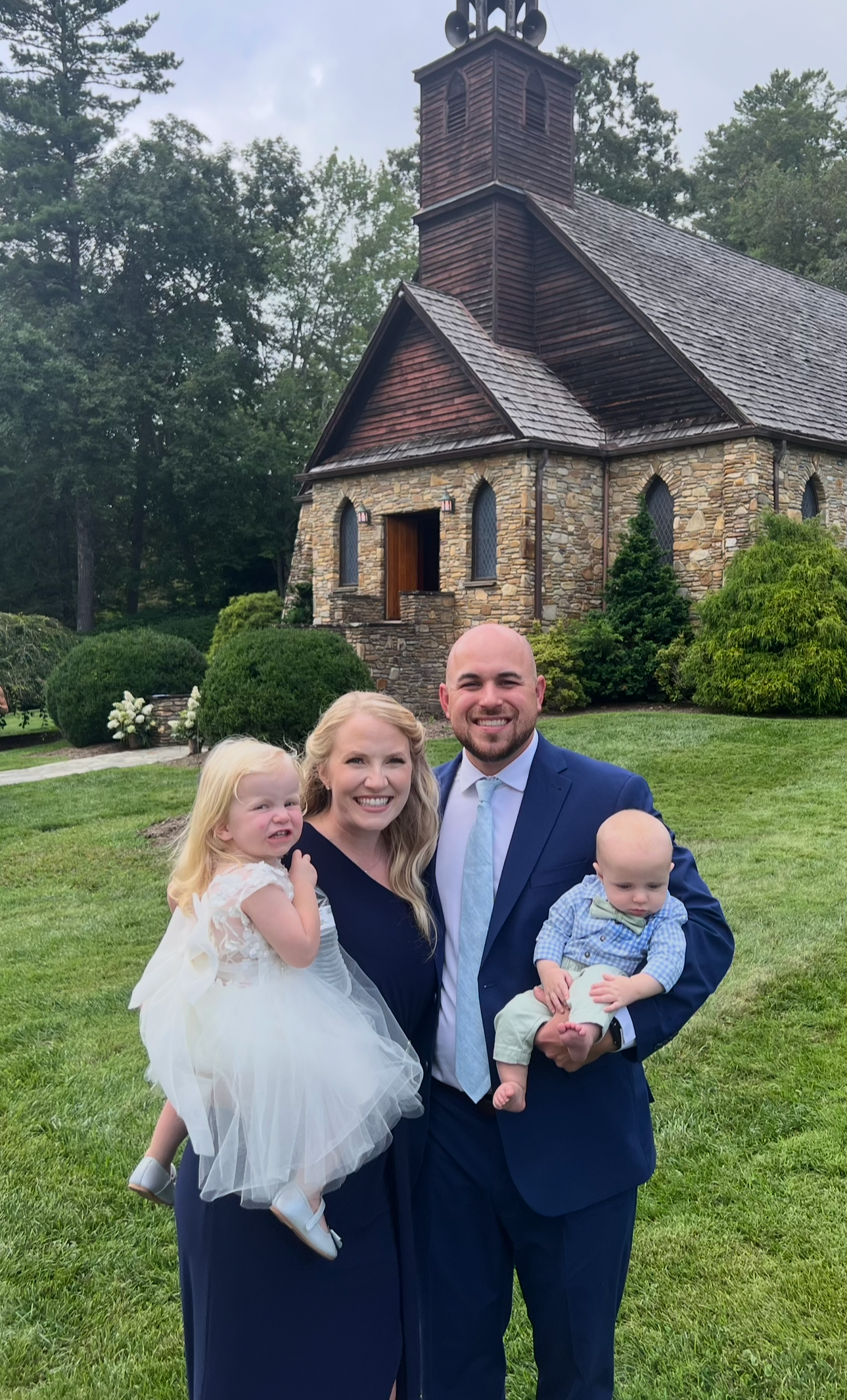 A family of four standing outdoors in front of a stone church with a wooden steeple. The family includes a woman with long blonde hair, a man in a suit, and two young children, a girl in a white dress and a boy in a blue shirt and bow tie. The woman is smiling and holding the girl, who appears to be crying or upset. The man is smiling and holding the boy.