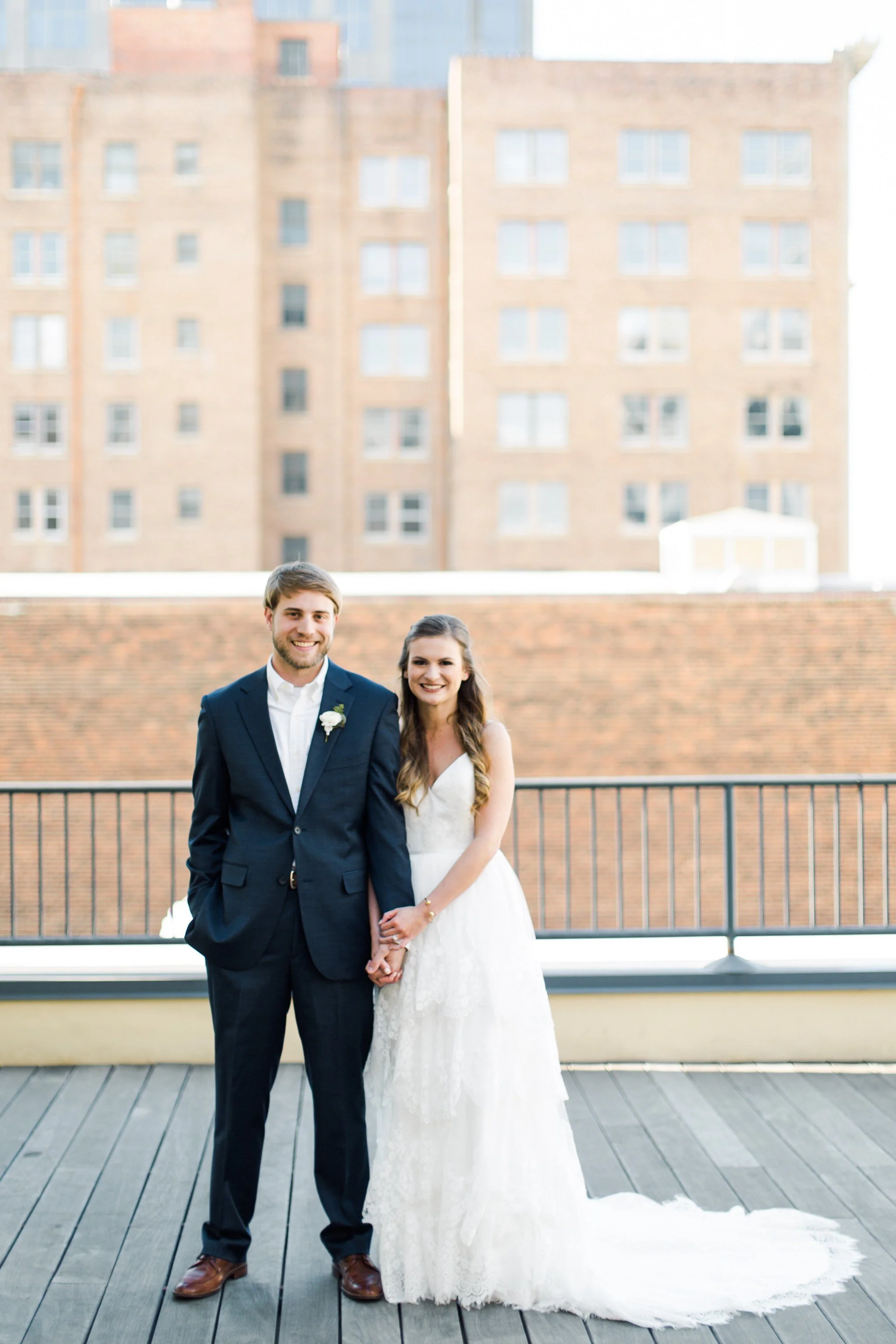 A newlywed couple standing hand in hand on a rooftop balcony with city buildings in the background, smiling at the camera.