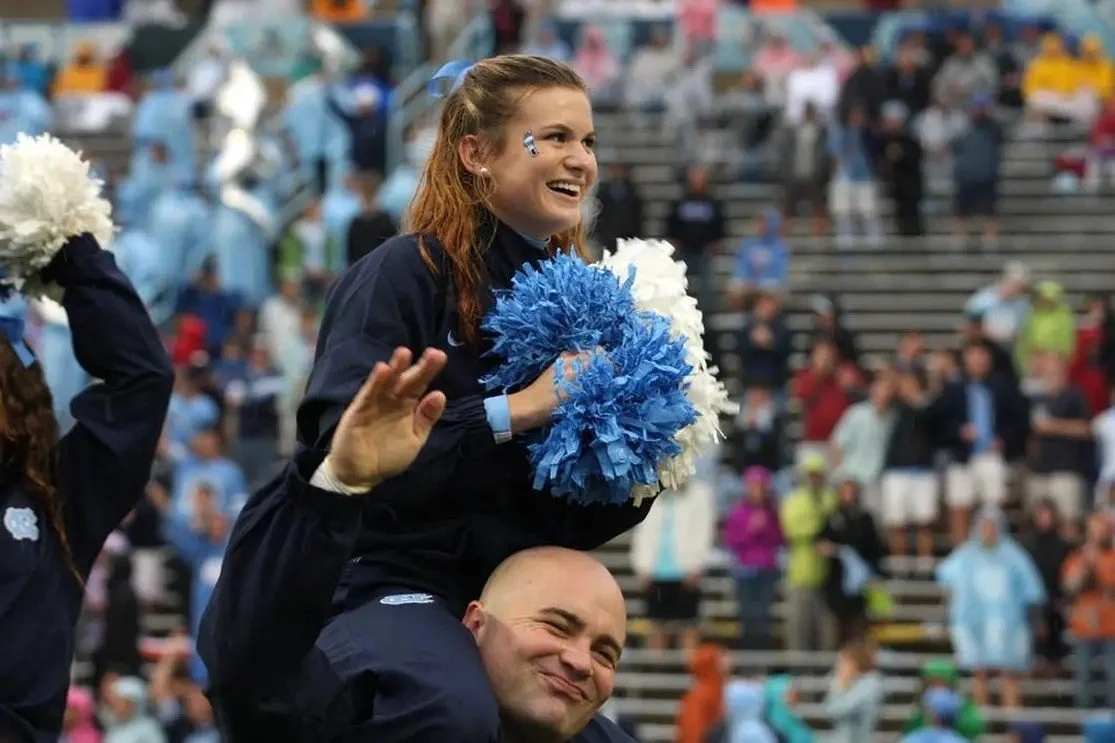 A young woman with red hair, wearing a navy jacket, is sitting on a man's shoulders at a sports event. She is smiling, holding blue and white pom-poms, and has a small sticker on her face. The background shows an audience in a stadium.