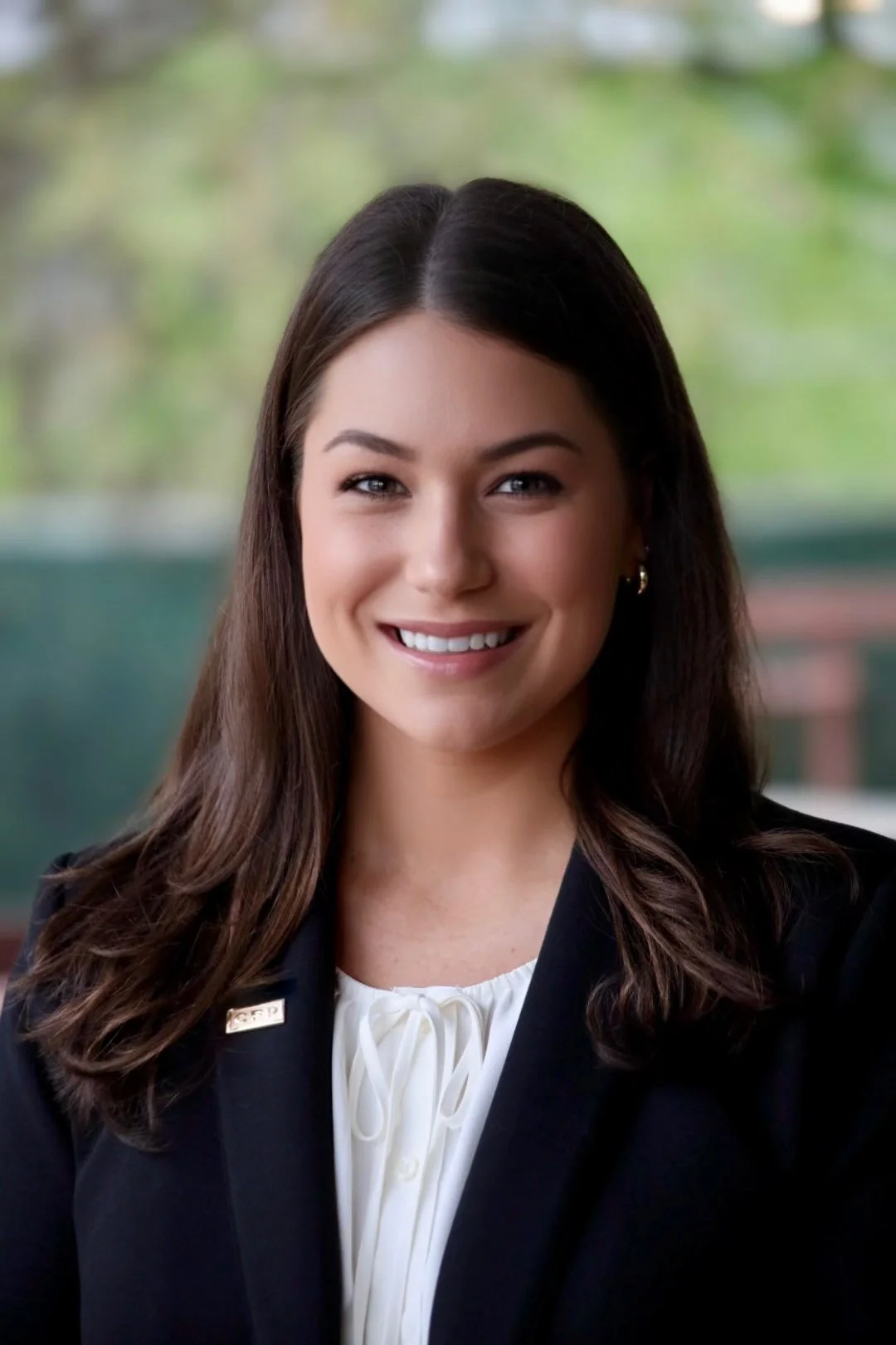 A woman with long brown hair, smiling, wearing a black blazer and a white blouse, with a blurred outdoor background.