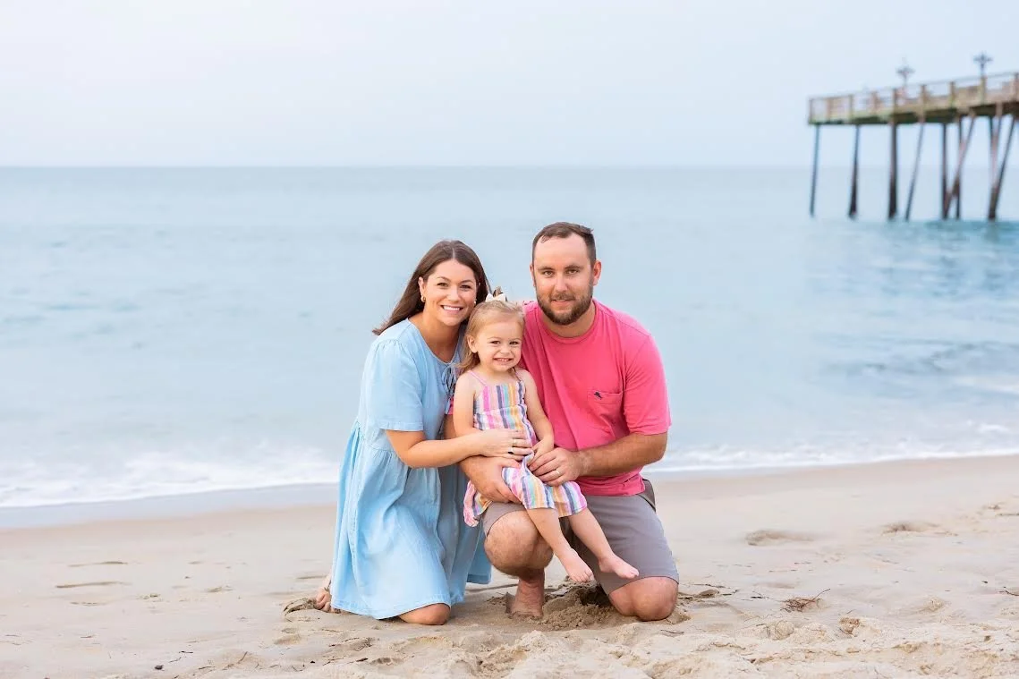 A family of three, consisting of a woman, a man, and a young girl, posing on a sandy beach with the ocean and a pier in the background.
