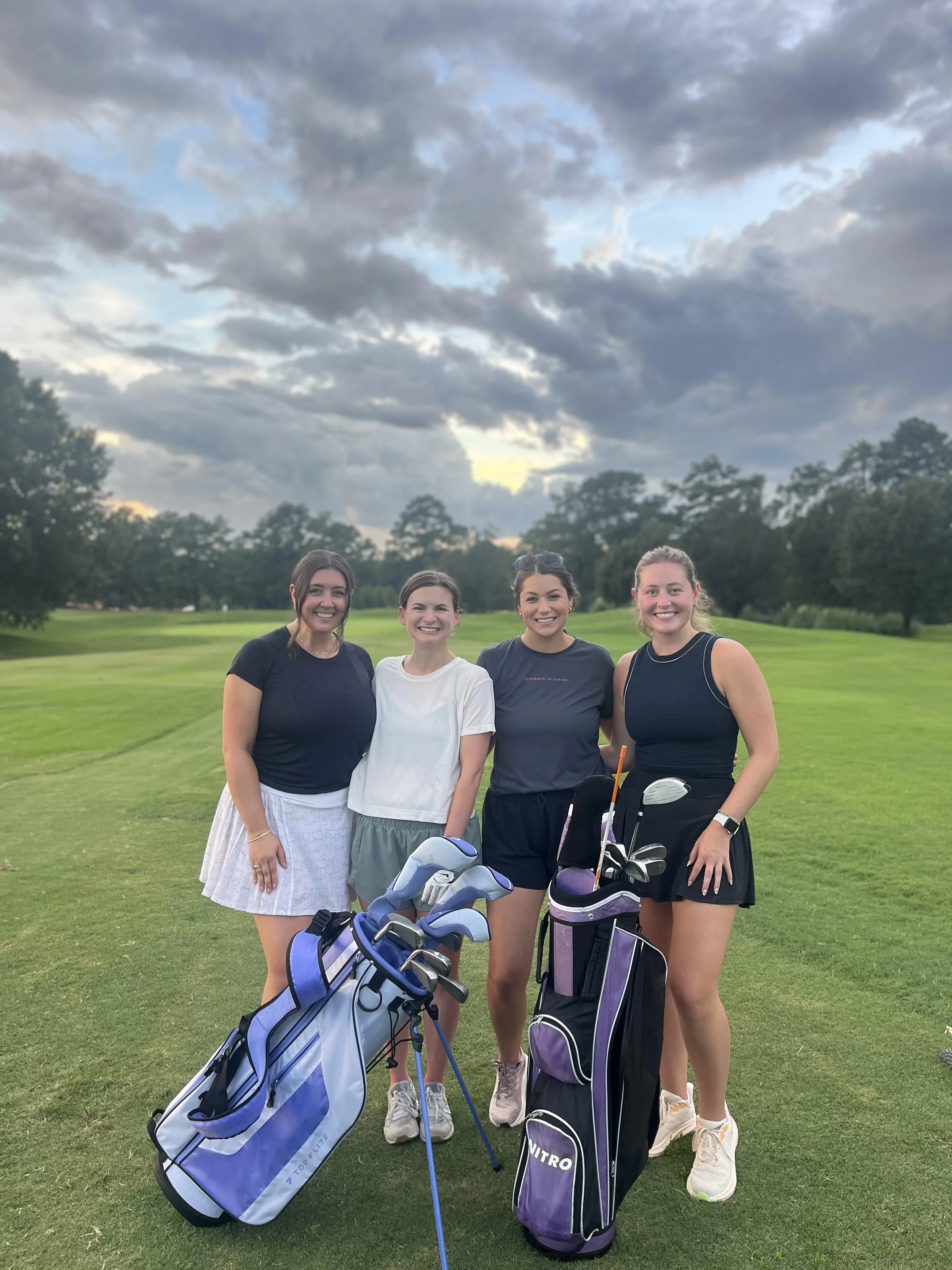 Four women standing on a golf course with golf bags and clubs, smiling at the camera, under a cloudy sky.