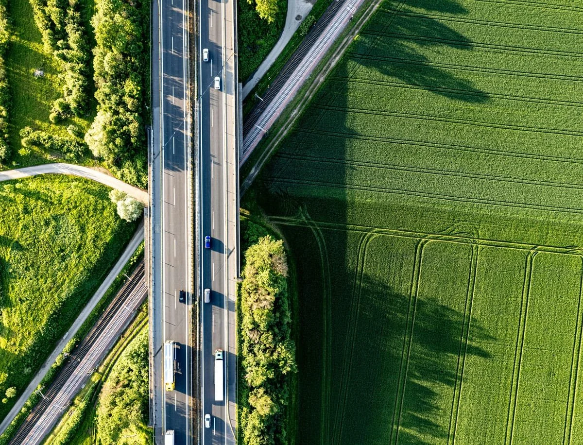 Aerial view of a highway passing through green farmland and forested areas, with train tracks running parallel beneath the highway.