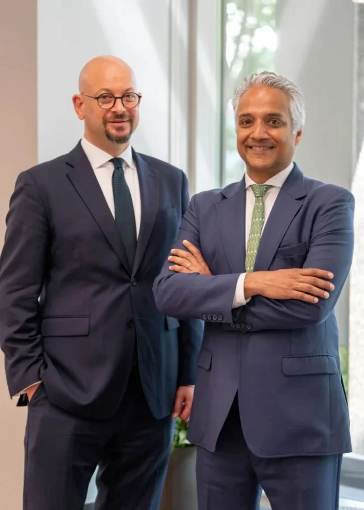 Two professionally dressed men in suits standing by a large window in an office, smiling at the camera.