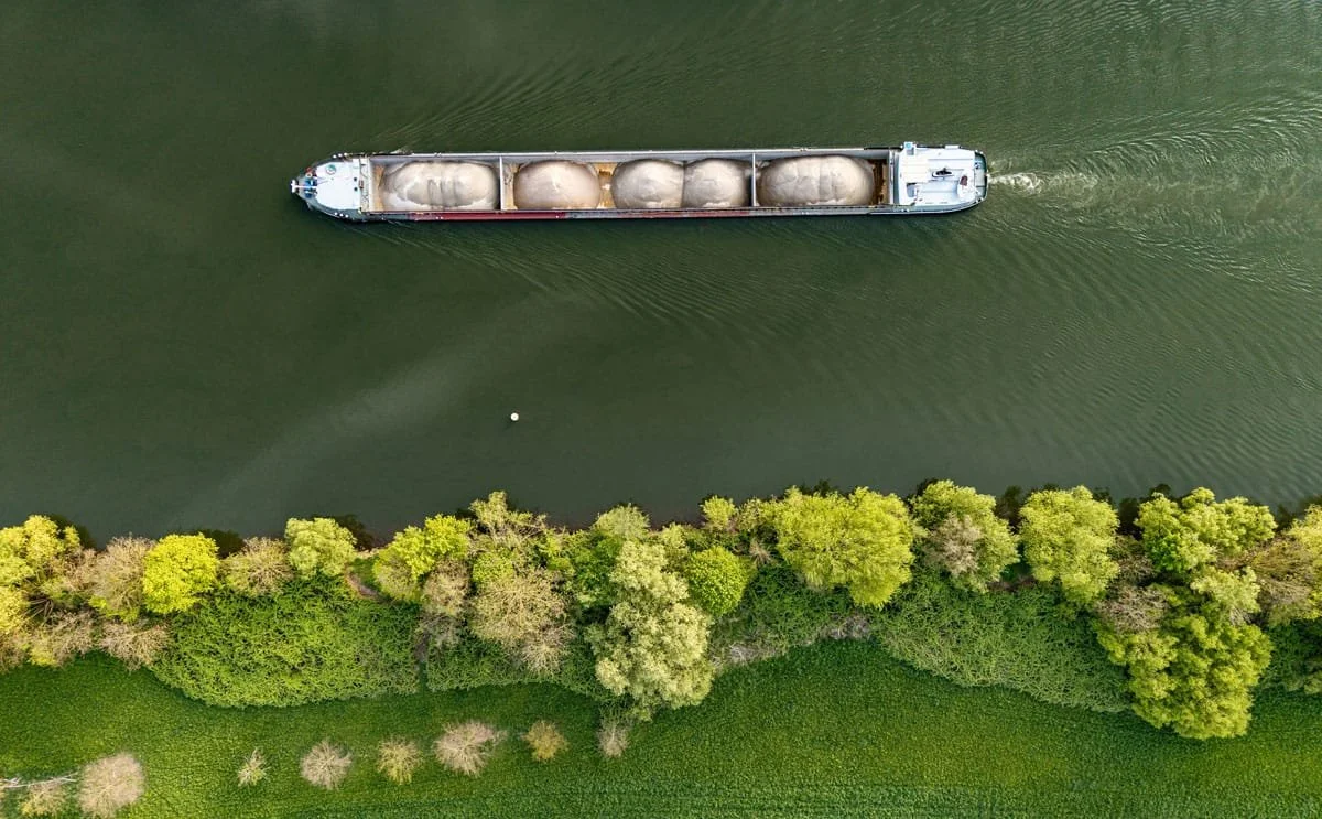 Aerial view of a cargo ship carrying large white dome-shaped containers on a river, with green trees and grass along the riverbank.