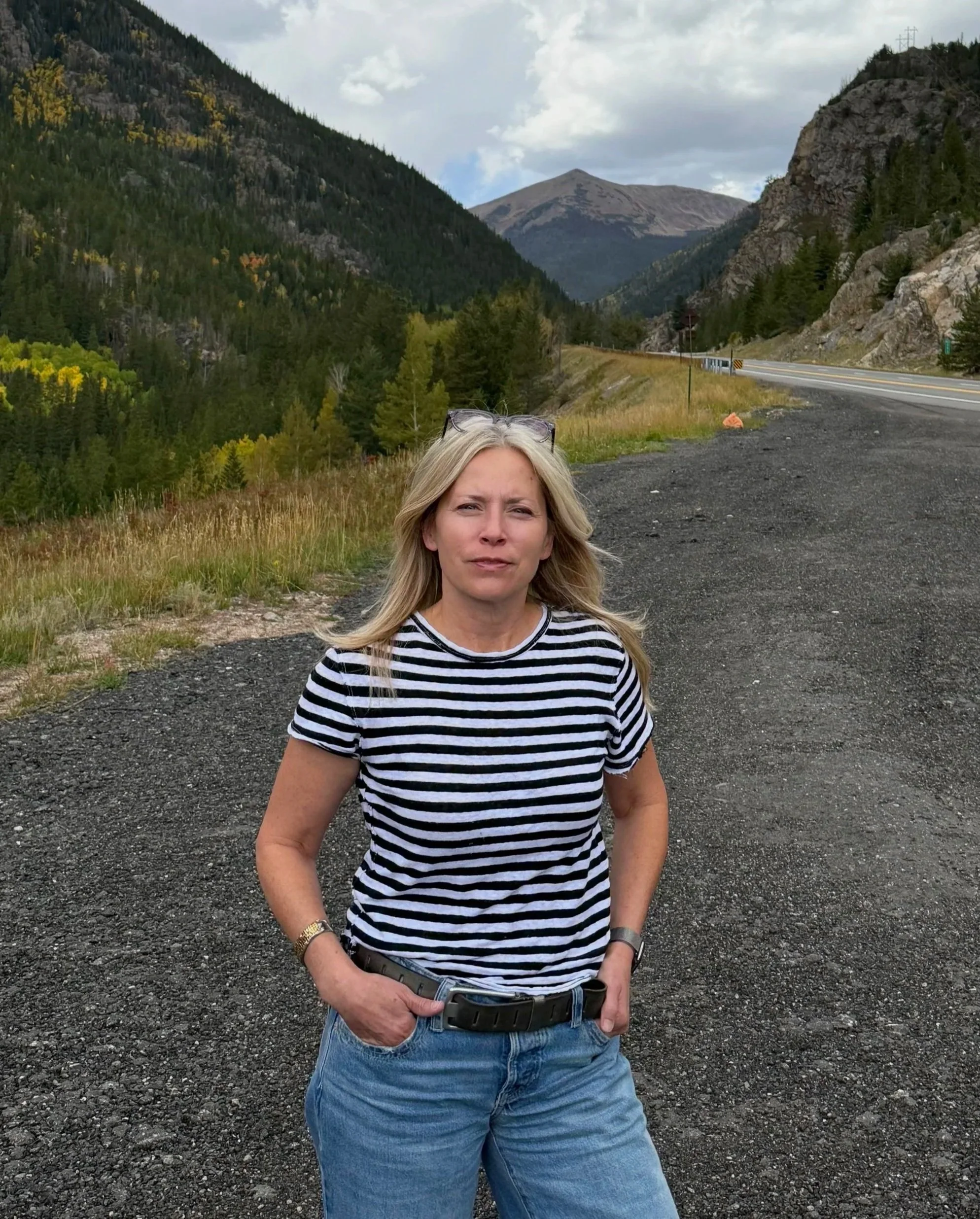 A woman with blonde hair wearing a striped shirt and jeans, standing on a gravel roadside in a mountainous area with Valley views, some trees, and a cloudy sky.