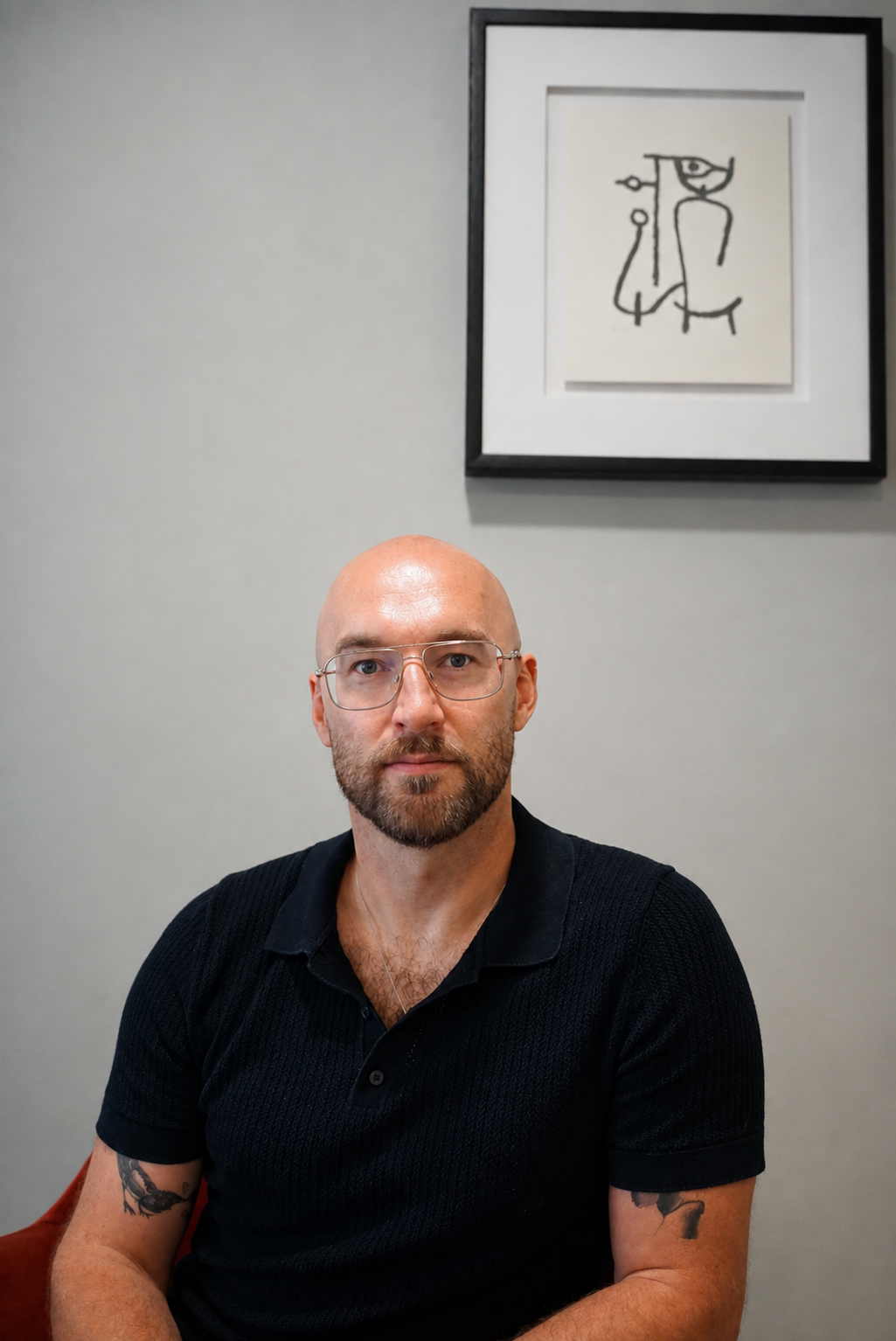 Adam Sweeney sitting in front of a gray wall, with a framed black and white abstract line drawing hanging above him.