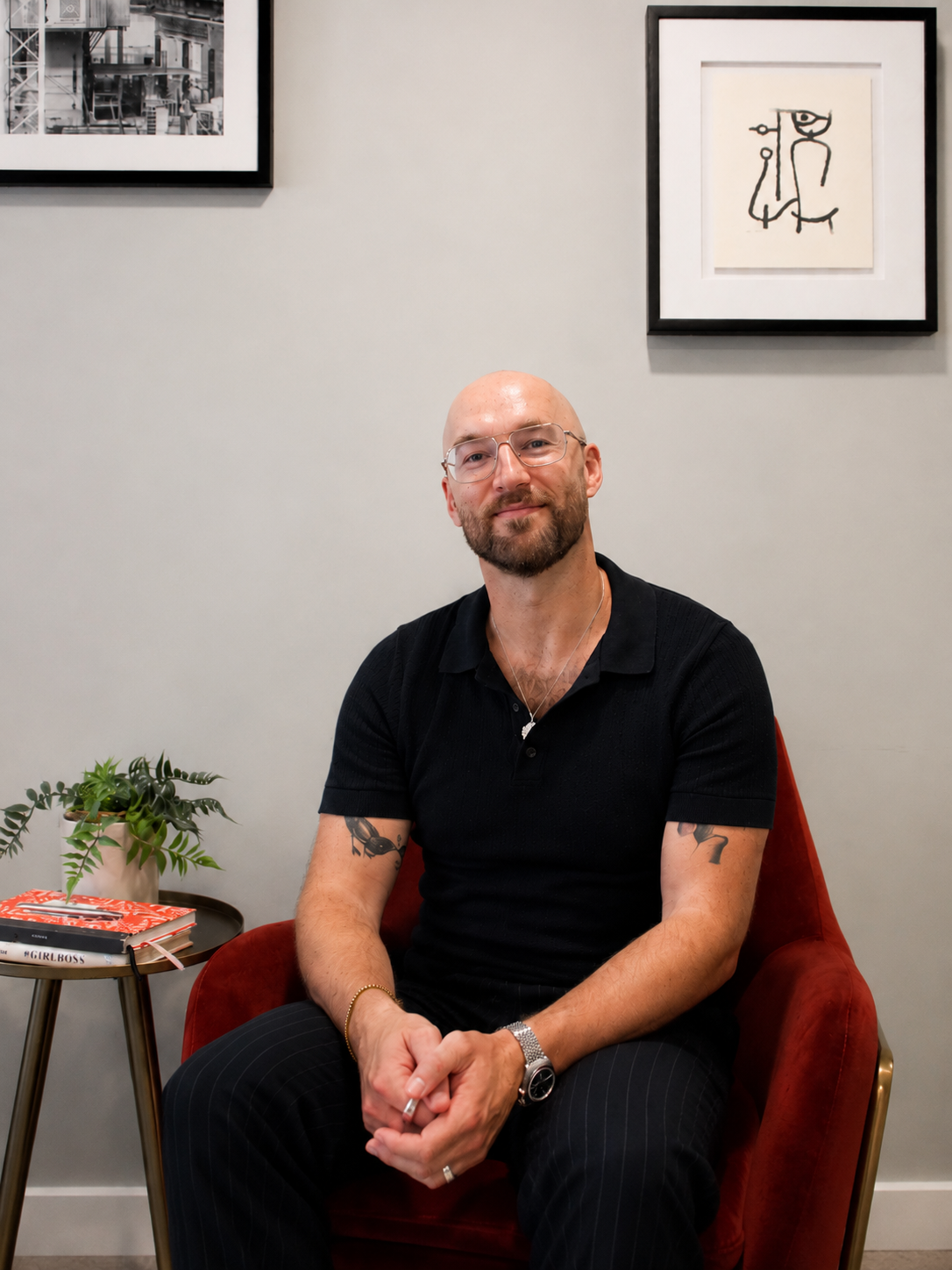 Adam Sweeney sitting in a red chair in front of a gray wall decorated with framed artwork. There is a small side table with books and a potted plant to his left.