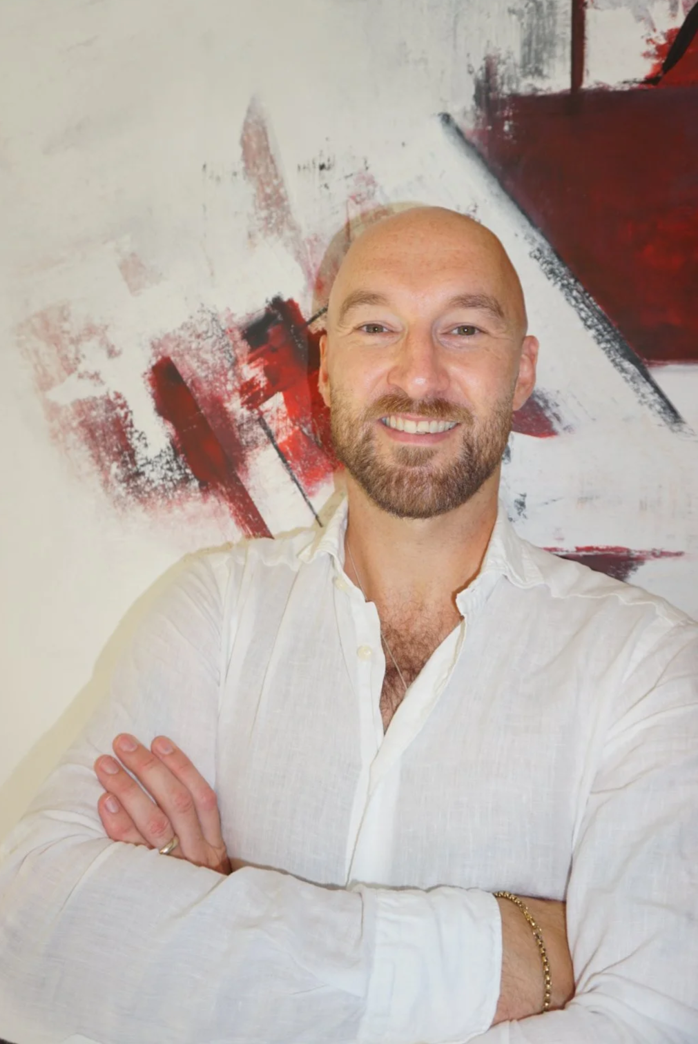 Adam Sweeney smiling, wearing a white shirt, standing in front of an abstract red and black painting.