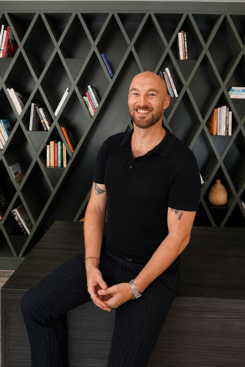 Adam Sweeney wearing a black polo shirt and striped pants, sitting on a dark wood bench in front of a black geometric bookshelf filled with books and decorative items.