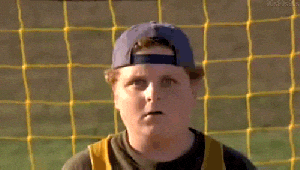 A young boy in a baseball uniform standing in front of a yellow net, wearing a backward cap, with an intense expression.