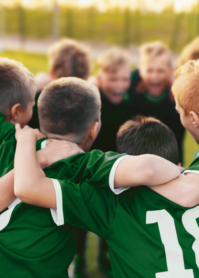 A group of young boys in green soccer uniforms huddled together in a circle outdoors, with hills and trees in the background.