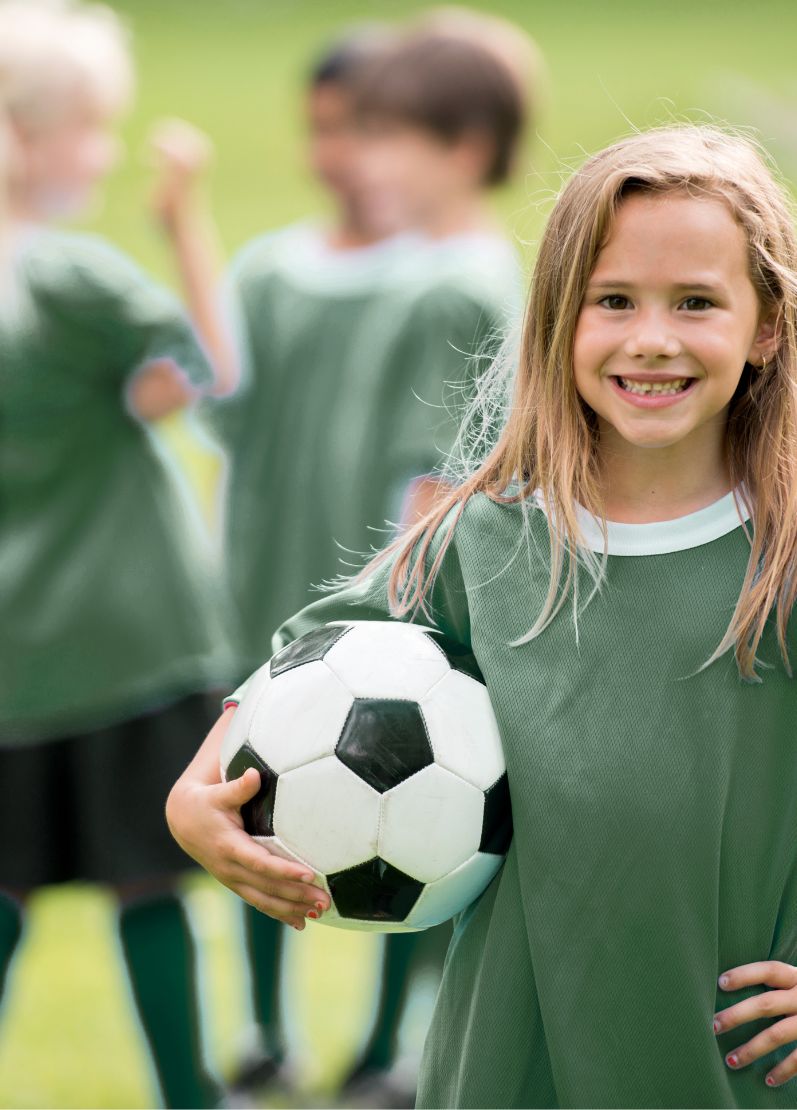 Young girl holding a soccer ball on a soccer field with other children playing in the background.