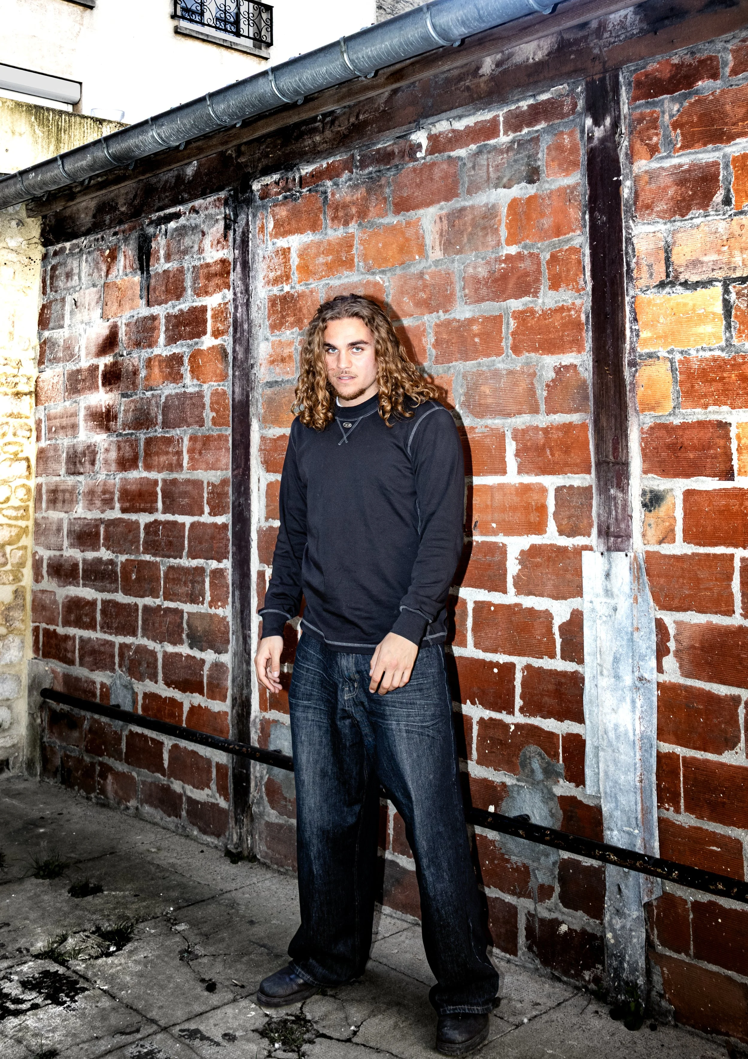 Jeune homme aux cheveux longs, portant un sweat noir et un jean ample, debout devant un mur en briques rouges dans une rue pavée.