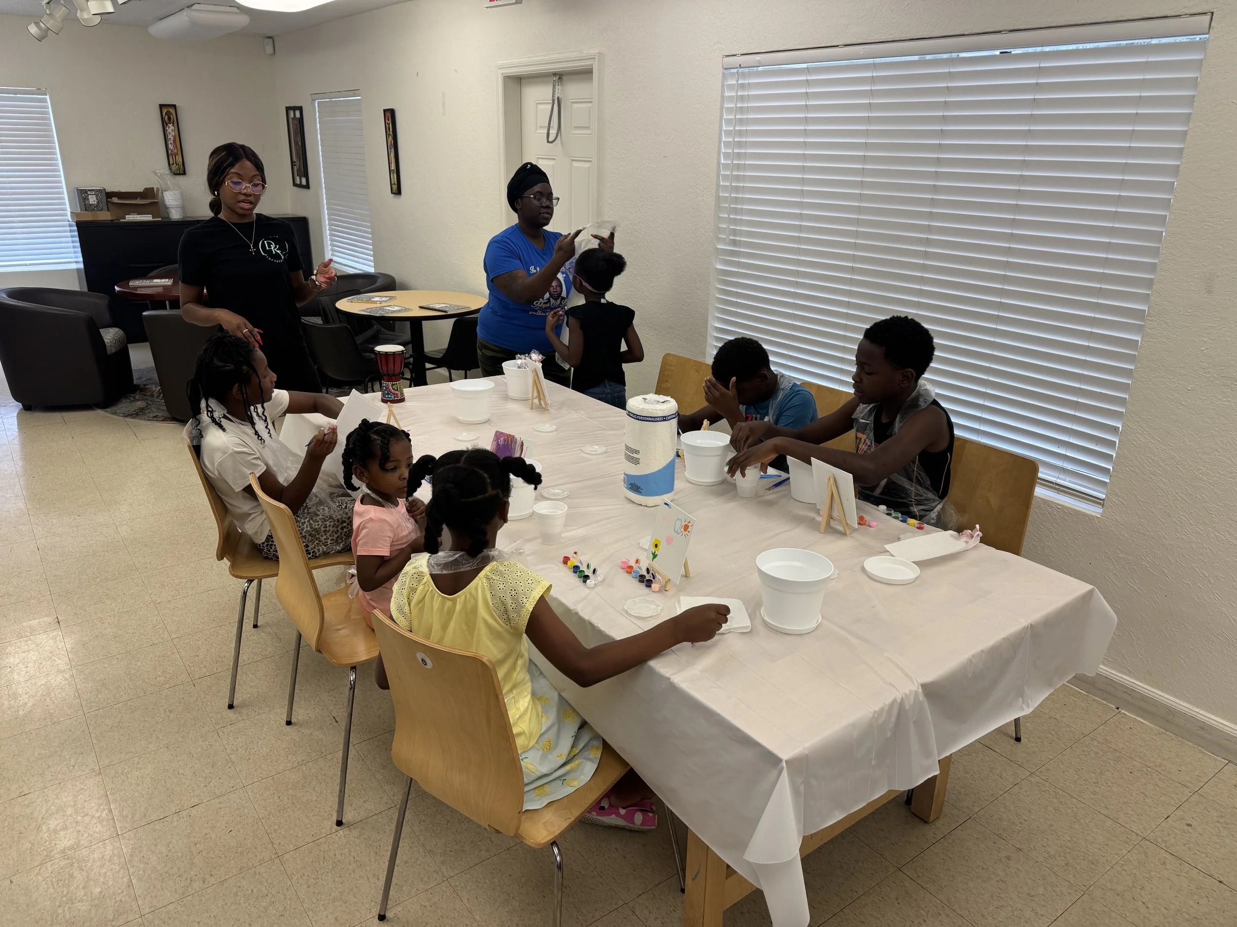 Children and adults participating in a craft activity at a table covered with a white tablecloth, with supplies like paint and cups, in a bright room with blinds covering windows.