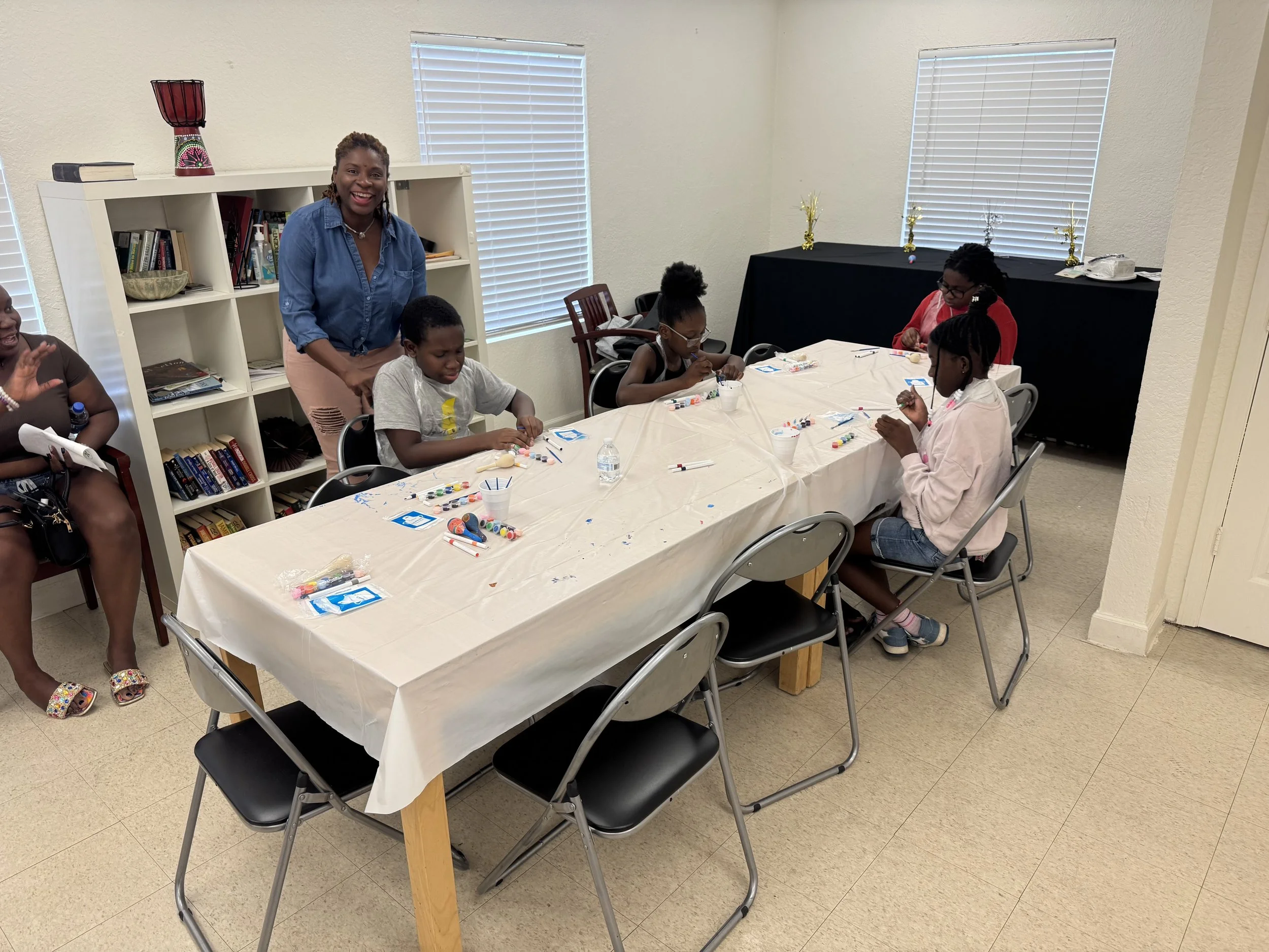 A group of children and an adultwoman gathered around a table with arts and crafts supplies in a bright room. The woman is smiling and standing behind the children. There are bookshelves, windows with blinds, and decorations in the background.