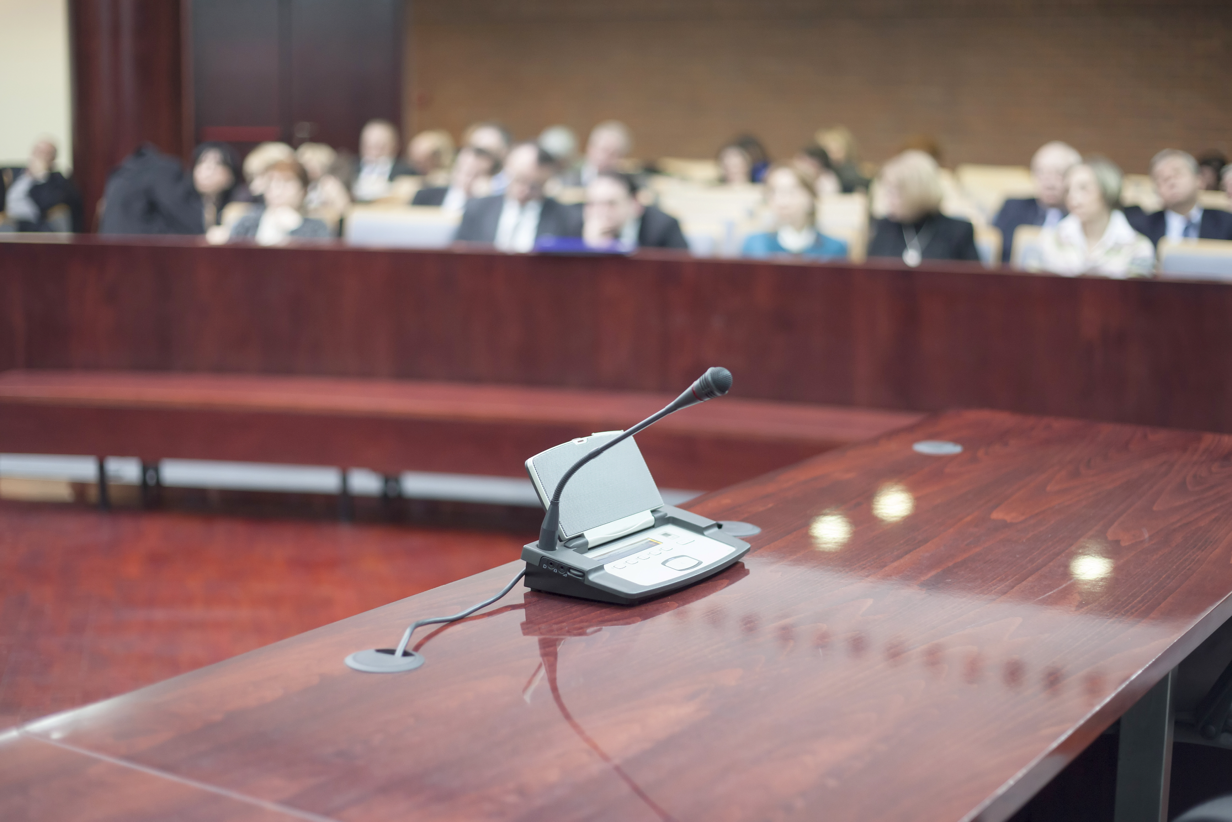 Empty conference room with a wooden table, microphone, and a listening device, with blurred attendees sitting in the background.