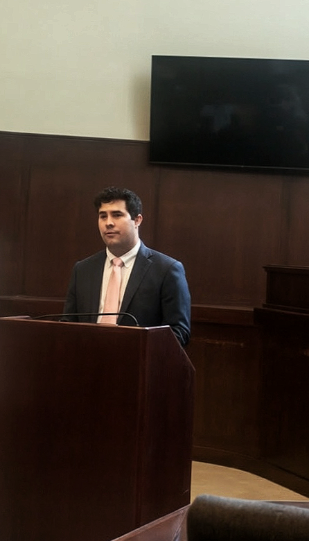 A man in a suit and tie standing at a wooden podium in a courtroom or legal setting.