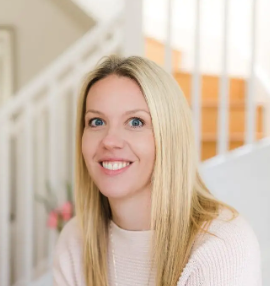 A woman with blonde hair and blue eyes smiling in a bright, indoor setting with a staircase in the background.