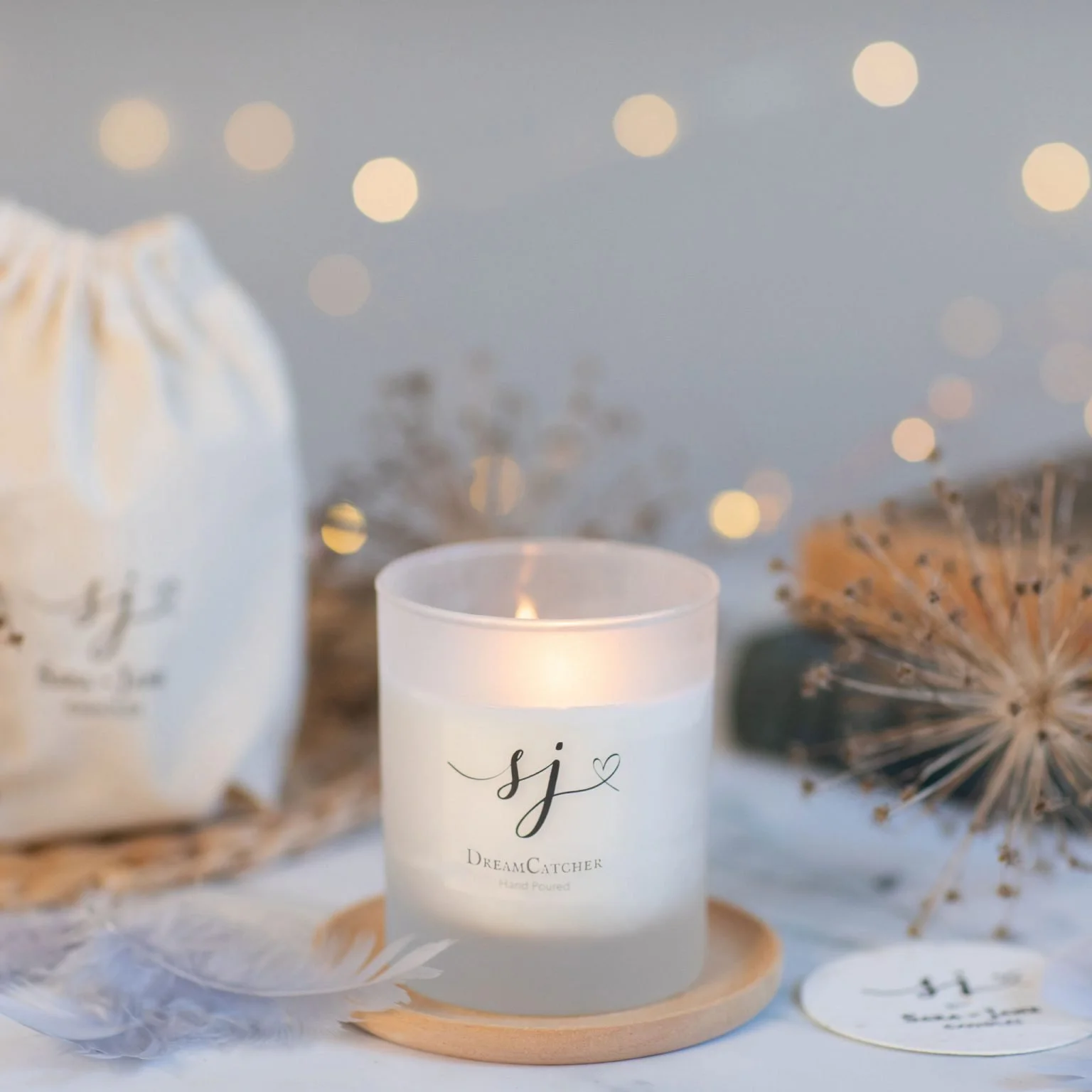 A lit white candle with the initials 'sj' and the words 'Dream Catcher Hand Poured' on a tray, with decorative dried plants and a white drawstring bag in the background with black text.
