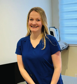 A young woman with blonde hair wearing blue scrubs, smiling indoors in front of a window with a white blinds and an electrical outlet.