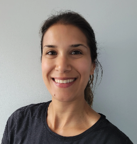 A smiling woman with dark hair tied back, wearing a dark T-shirt, standing against a light-colored wall.