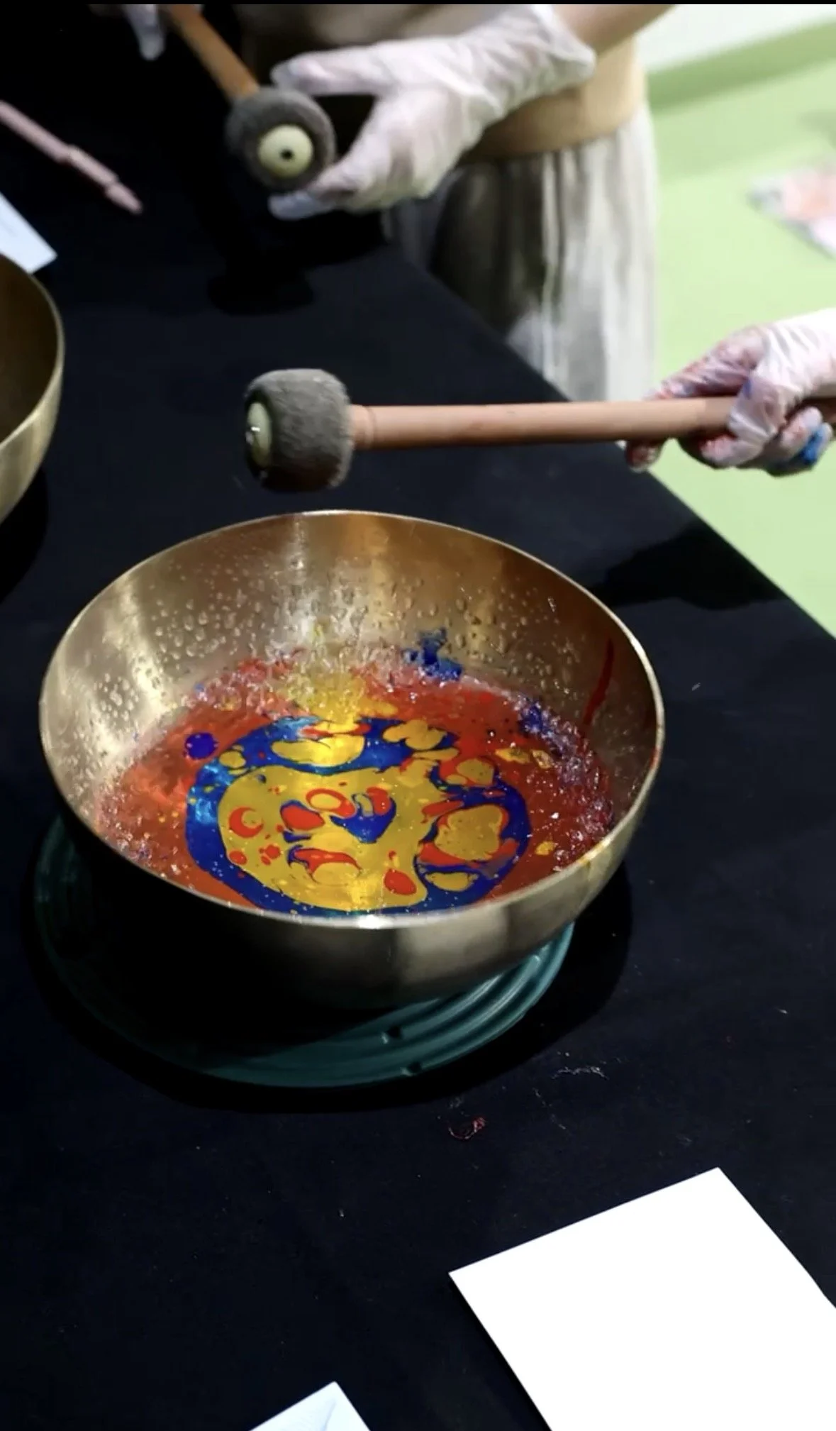A person wearing gloves is using a mallet to hit a singing bowl filled with colored liquid on a black table.
