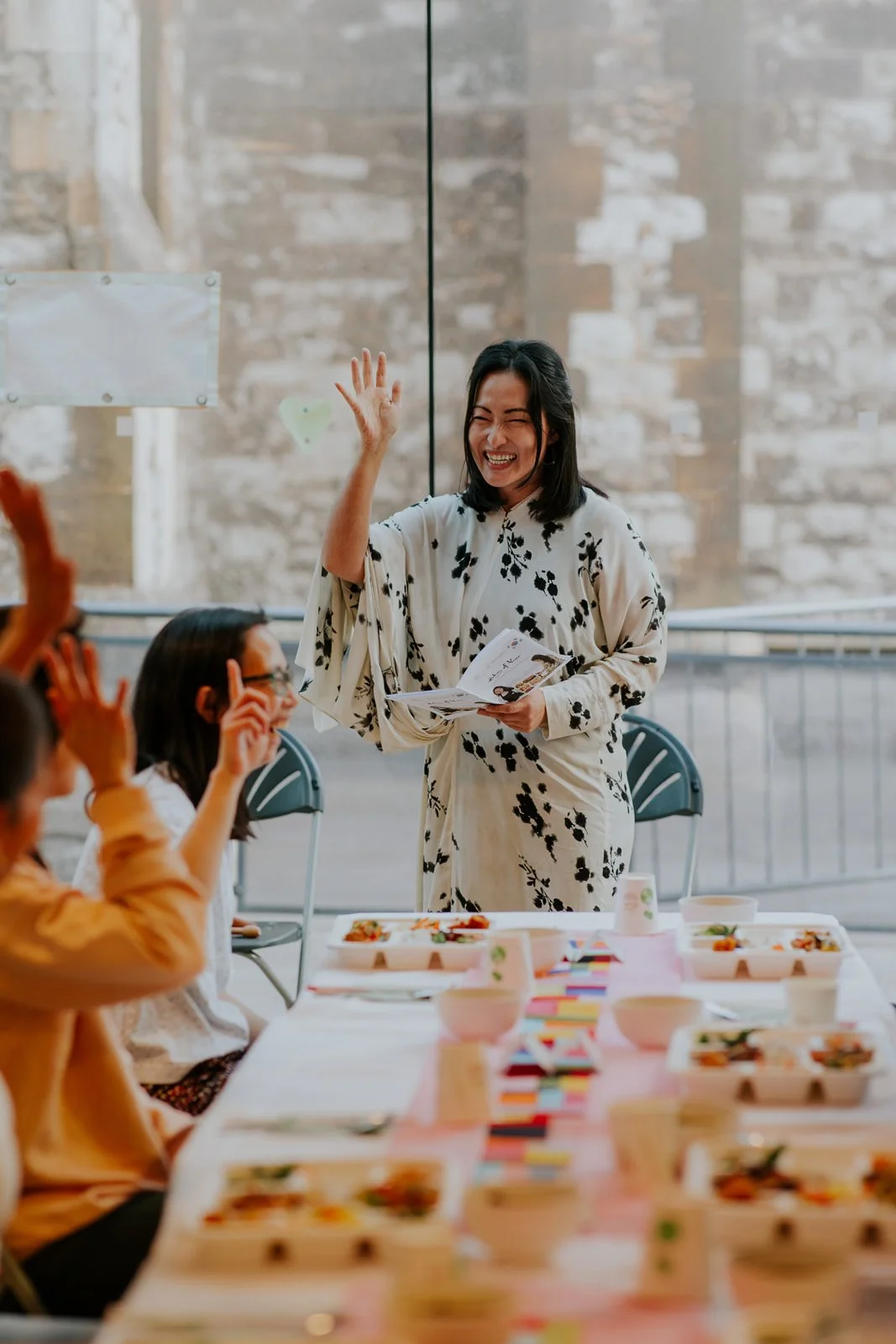 A woman standing and smiling, holding papers, raising her hand, in front of seated children at a table with food.