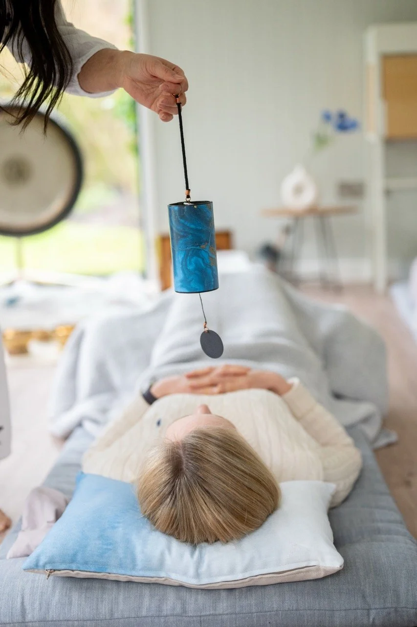 Person lying on a bed with a blue pillow, receiving an energy healing session with a practitioner holding a cylindrical device with a cord and a small disc at the end, above the person's forehead.