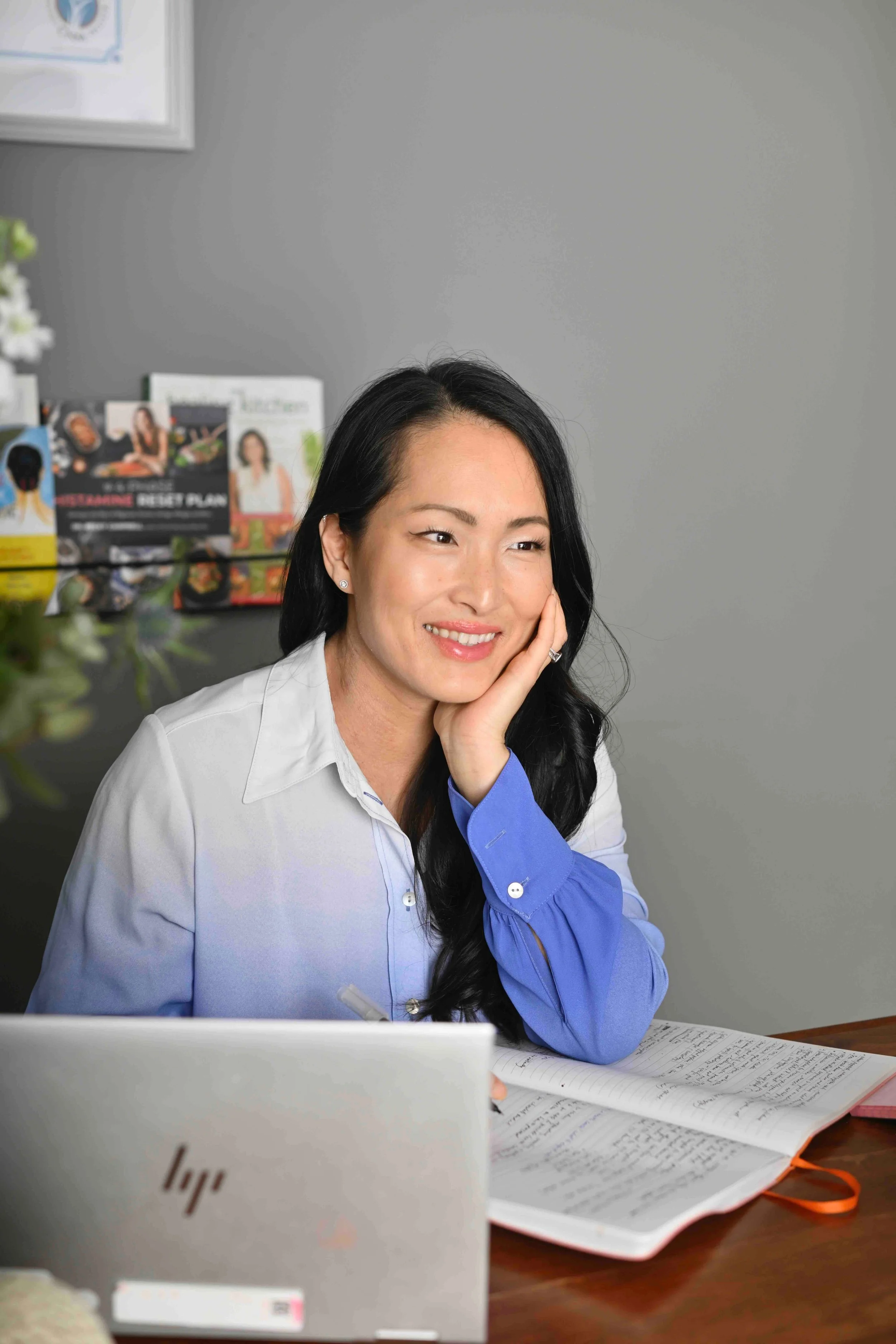 A woman with long black hair, wearing a blue and white shirt, sitting at a desk with an open notebook, a laptop, and some papers. She is smiling and resting her chin on her hand, looking to her left. The background shows a gray wall with a bulletin board and some magazines.