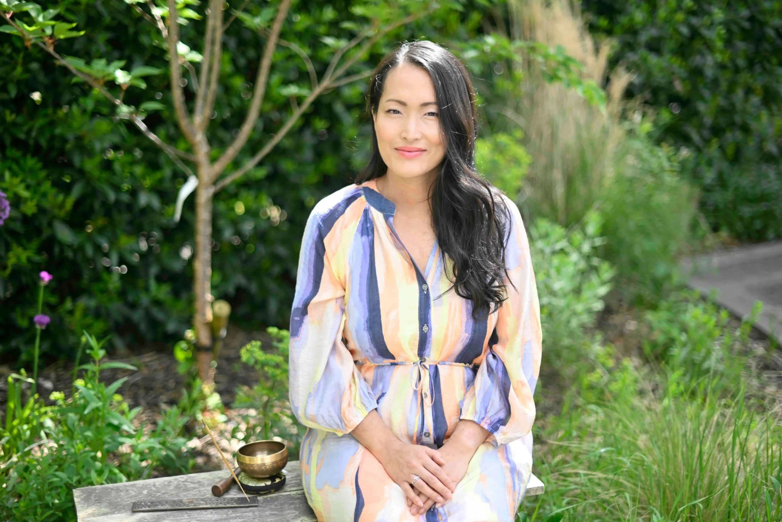 A woman with long dark hair wearing a colorful striped dress, sitting outdoors surrounded by lush green bushes and trees. There is a singing bowl in front of her on a wooden surface.