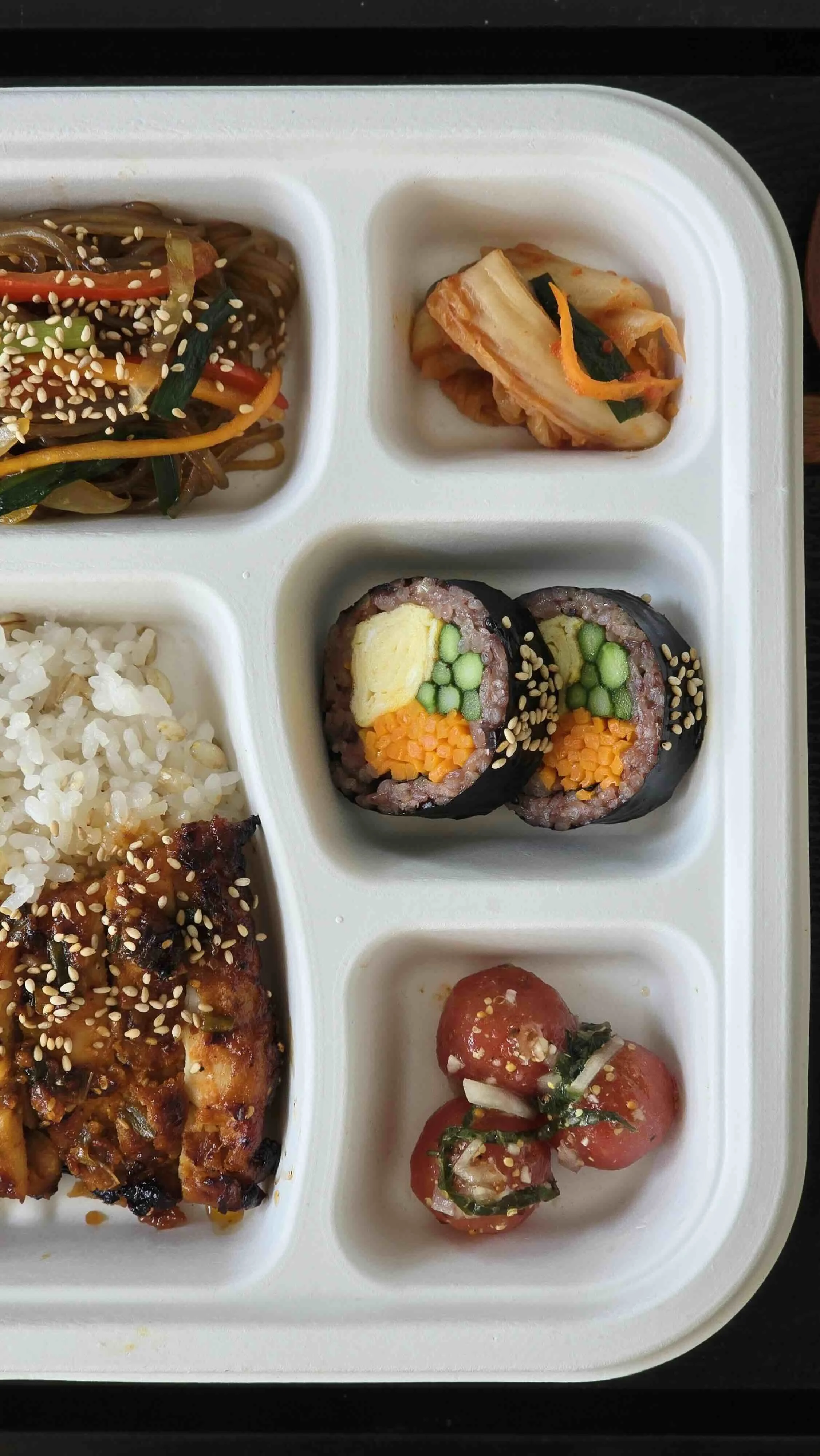A compartmentalized tray with Asian dishes including cold noodles with vegetables and sesame seeds, kimchi, rice with toppings, sushi rolls, and cherry tomatoes.