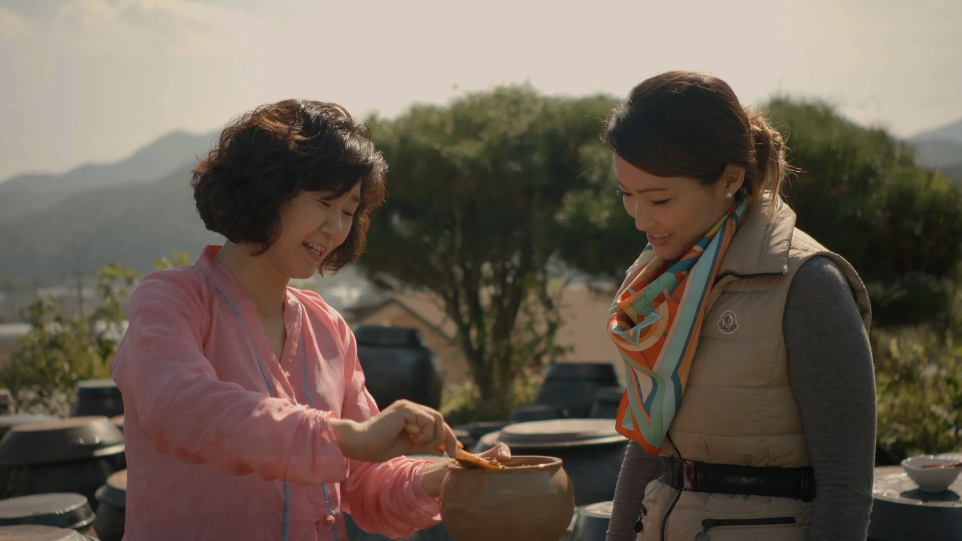 Two women smiling and engaging in a conversation outdoors, with pottery and trees in the background.