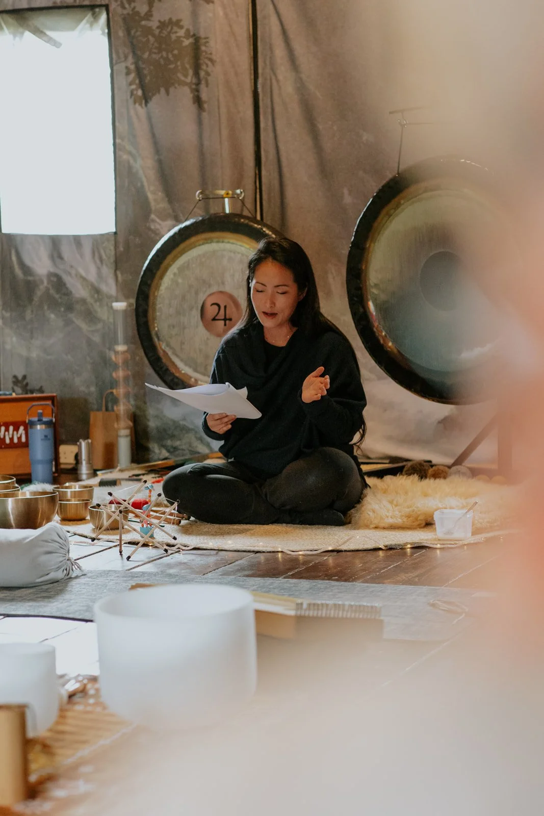 Woman sitting on rug in front of large gongs, holding papers and speaking, with bowls and musical instruments around her, in a cozy interior space.