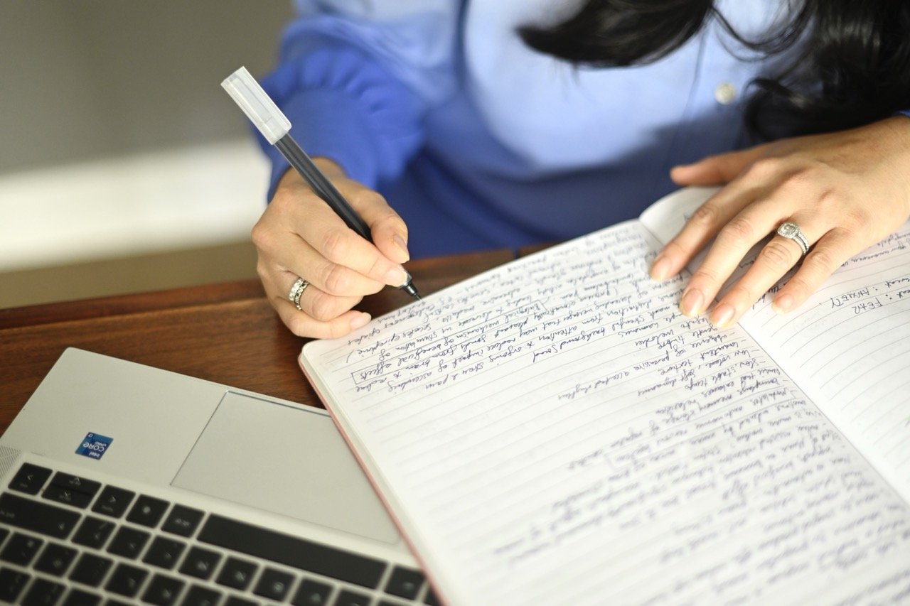 A person in a blue shirt writing in a notebook with a black marker, resting their hand on the open pages, with a silver laptop nearby.