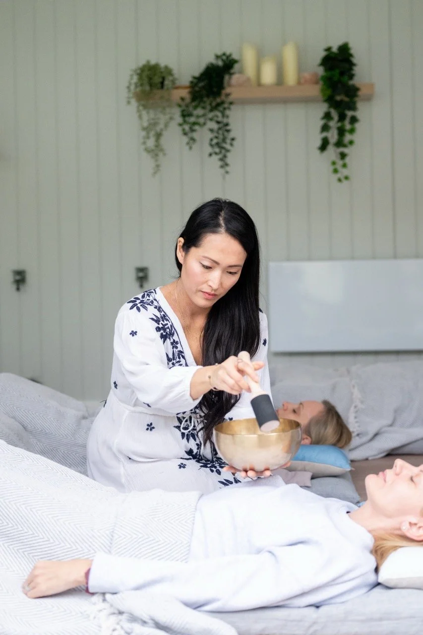 A woman performing sound therapy or sound healing session using a singing bowl on a person lying on a bed, with a meditation or yoga setting in the background.