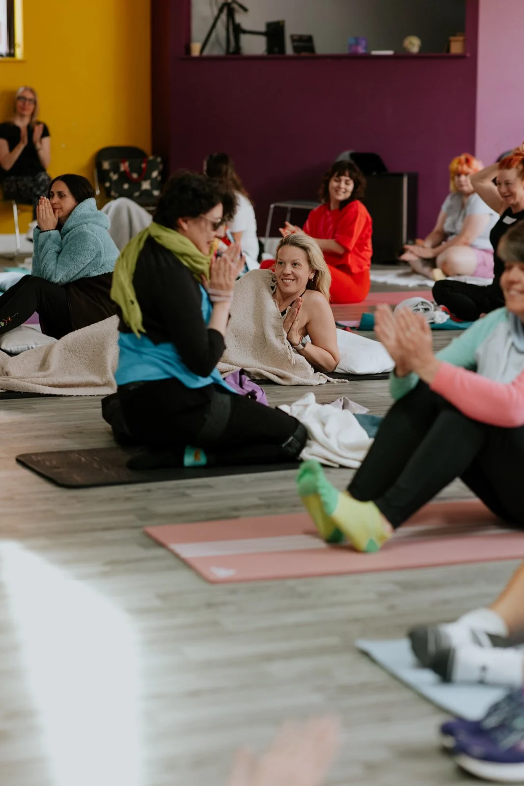 Group of women participating in a yoga or meditation class, sitting on mats on the floor, some smiling and engaging with each other in a brightly colored room.