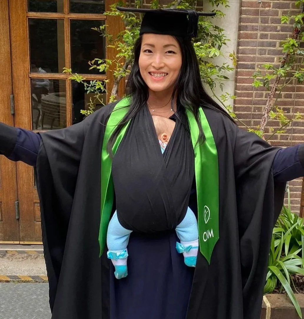 A woman in a graduation cap and gown with a newborn baby in a baby carrier in front, outdoors near a brick wall and greenery, celebrating graduation.