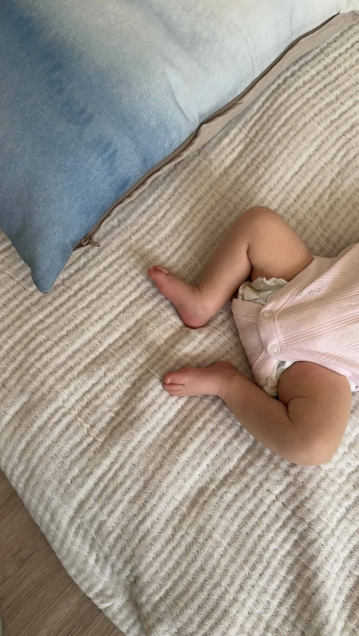 Close-up of a baby lying on a textured beige blanket on a bed, with a blue pillow and a pink onesie.