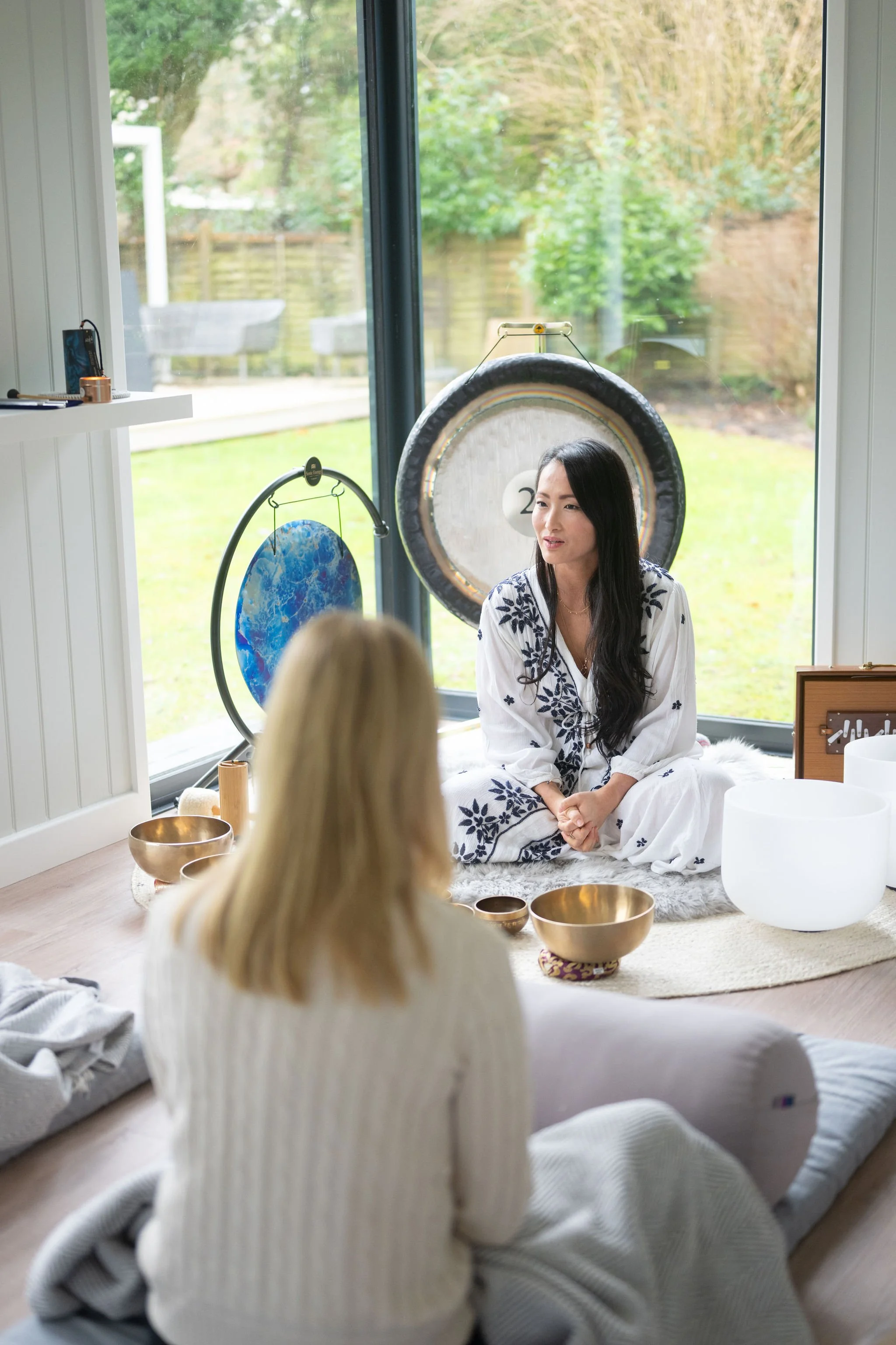 A woman with long dark hair, wearing a white dress with blue embroidery, sitting on a plush rug inside a room with large windows, a view of green outdoor foliage, and various singing bowls around her, participating in a meditation or sound healing session with a person in the foreground with blonde hair.